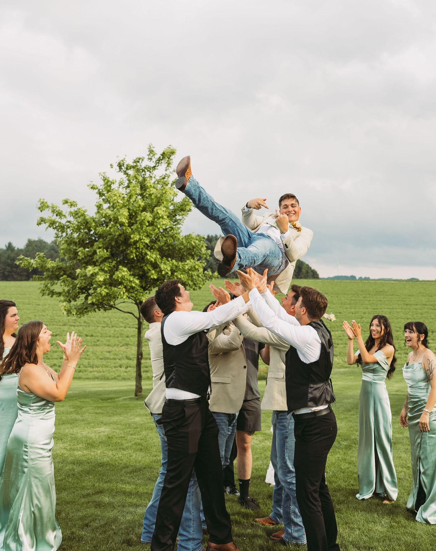 Wedding party celebrating outdoors by tossing someone in the air on a grassy field.