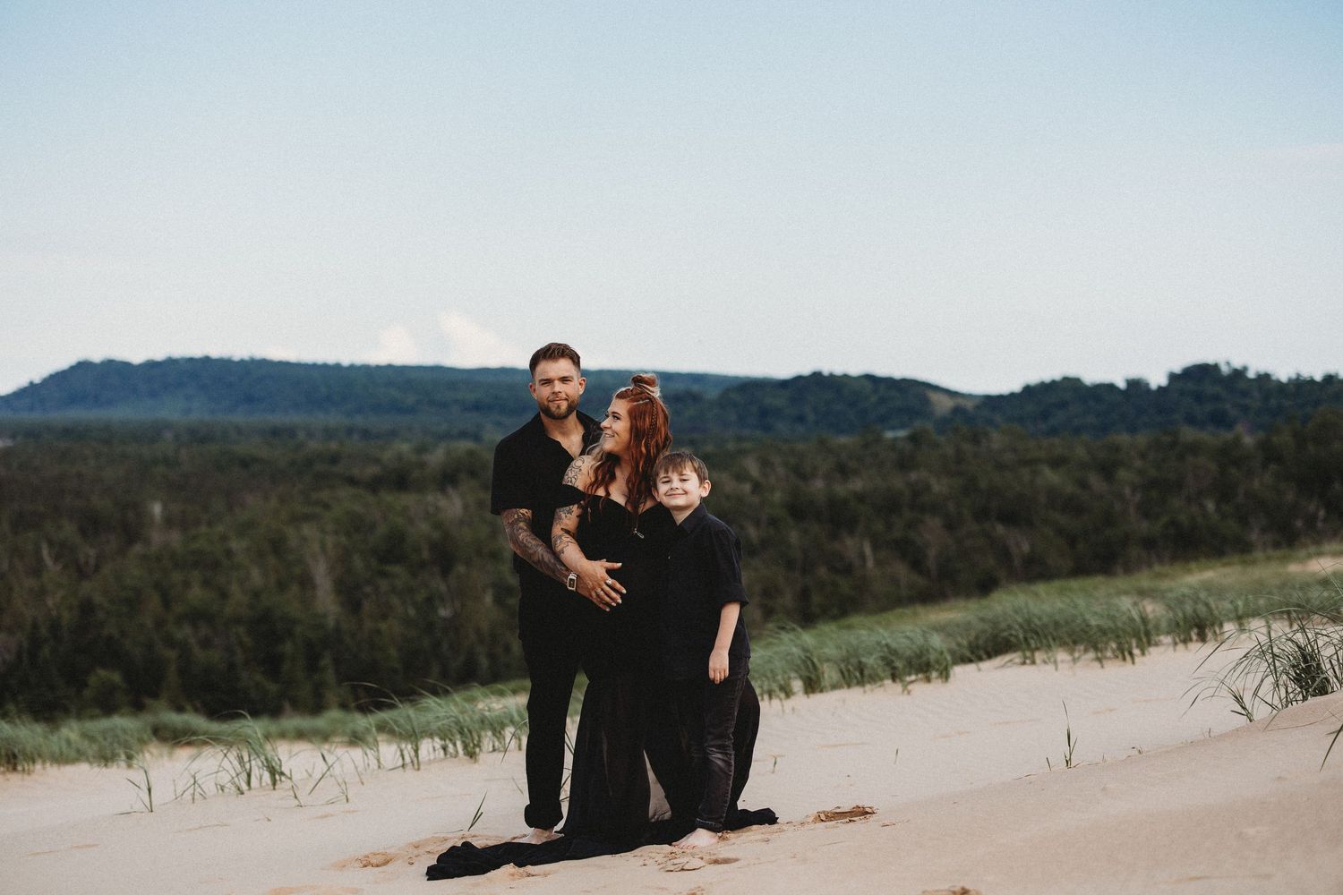 Two people stand together on sandy dunes with rolling hills and trees in the background on a bright day.