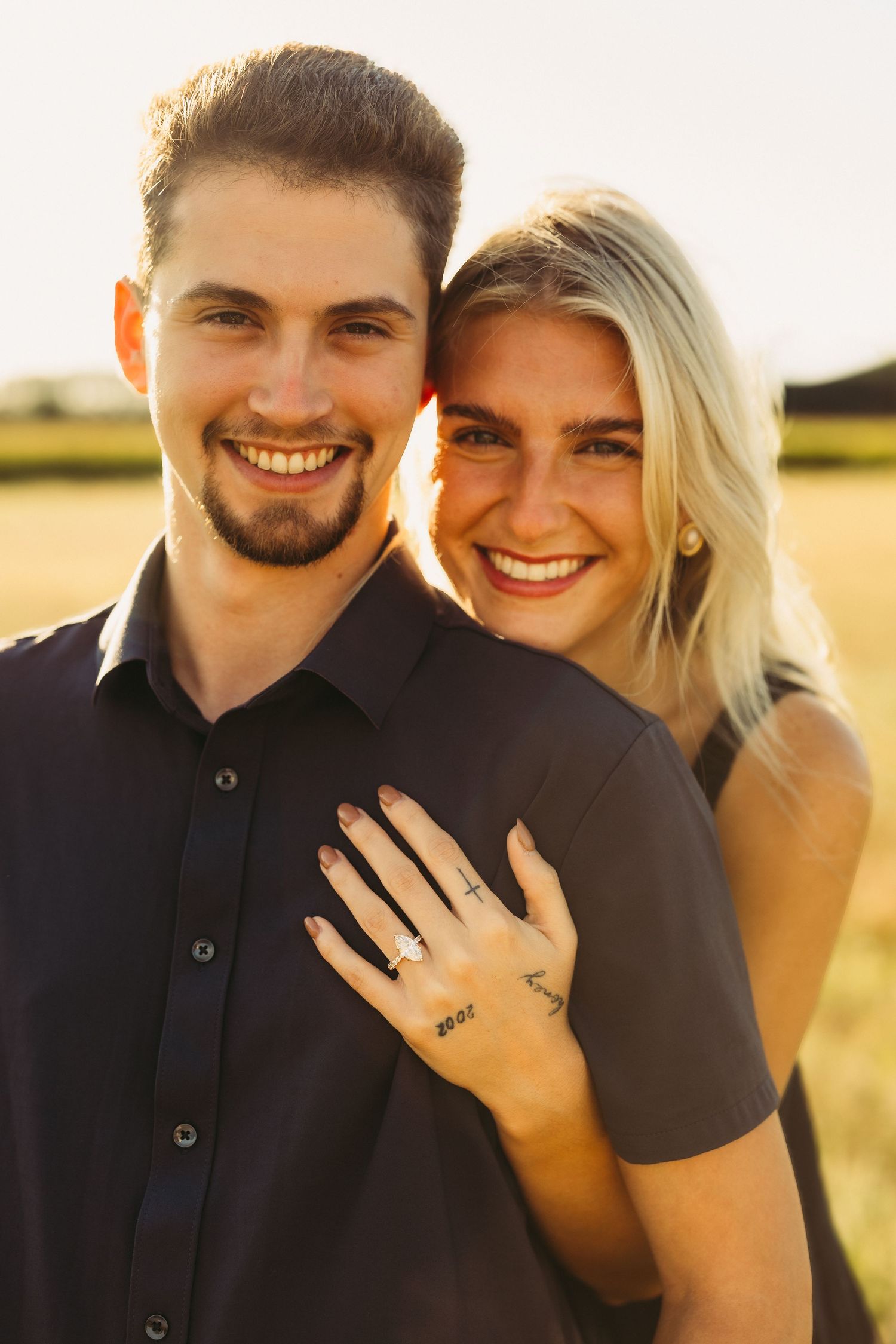 Smiling couple embracing in a golden field at sunset, with her ringed hand resting on his chest in an engagement photo.