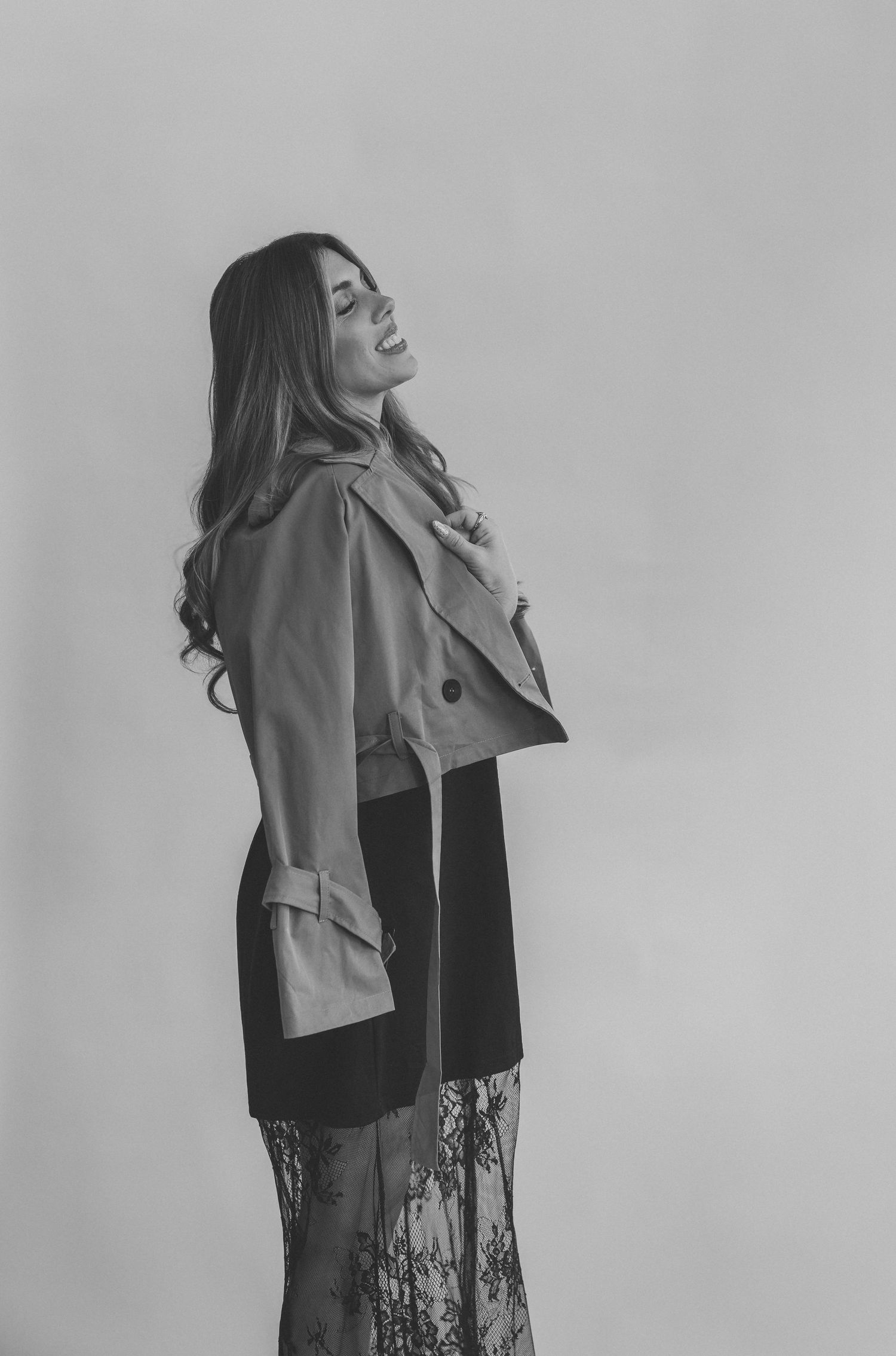 Black and white portrait of a laughing woman with long wavy hair wearing a cropped trench jacket over a lace skirt in a studio.