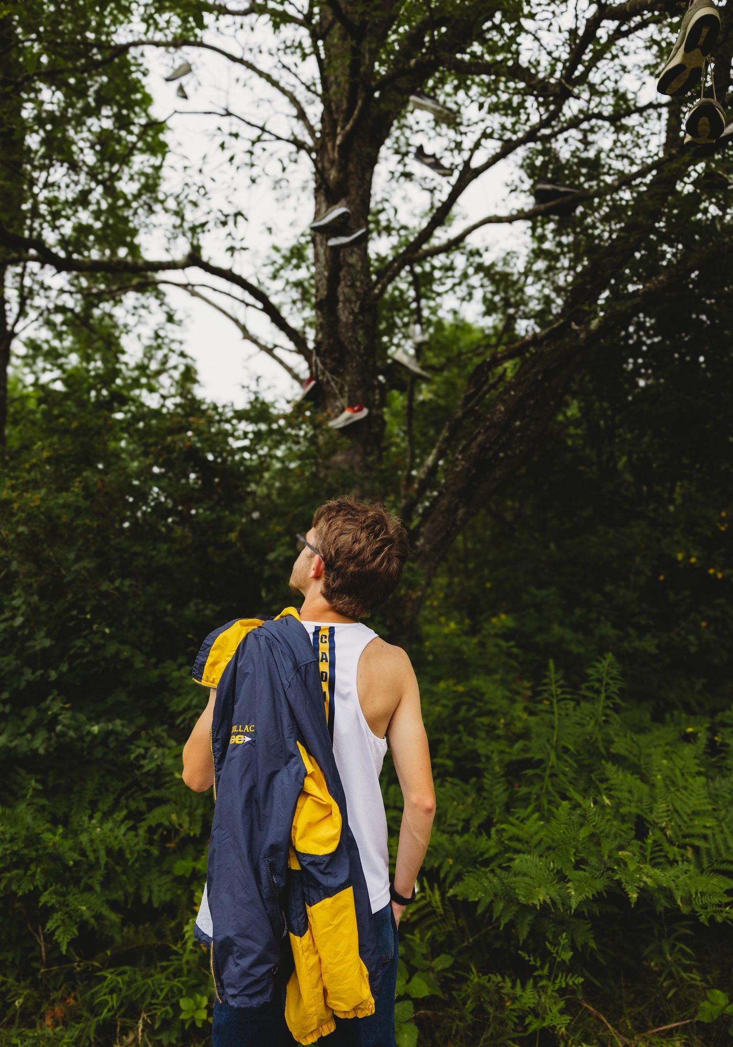 Person in a white tank top holding a navy and yellow jacket looks up at shoes hanging from tree branches in a lush green forest.