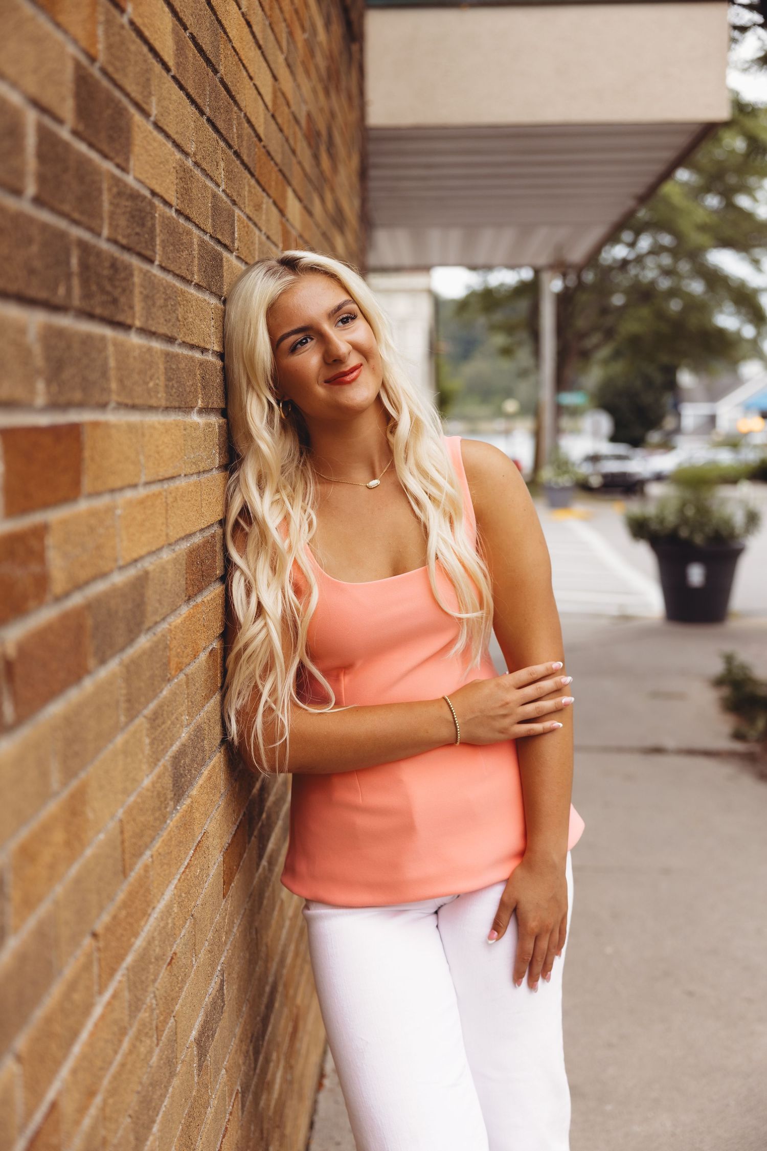 A smiling woman with long wavy blonde hair leans against a brick wall, wearing a coral tank top and white pants on a city sidewalk.