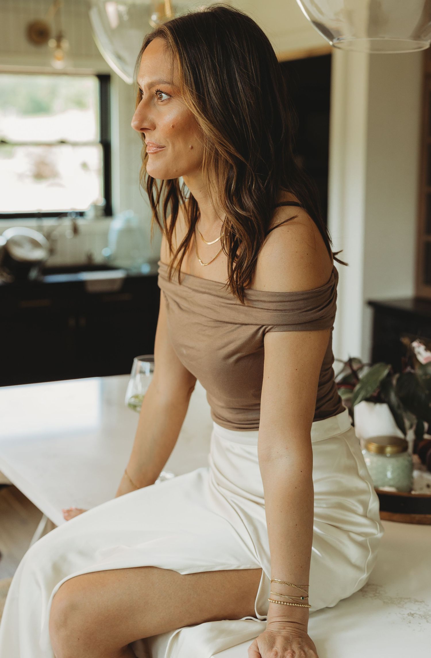 Woman in brown off-shoulder top and white skirt sitting on counter, wearing gold jewelry in modern kitchen.