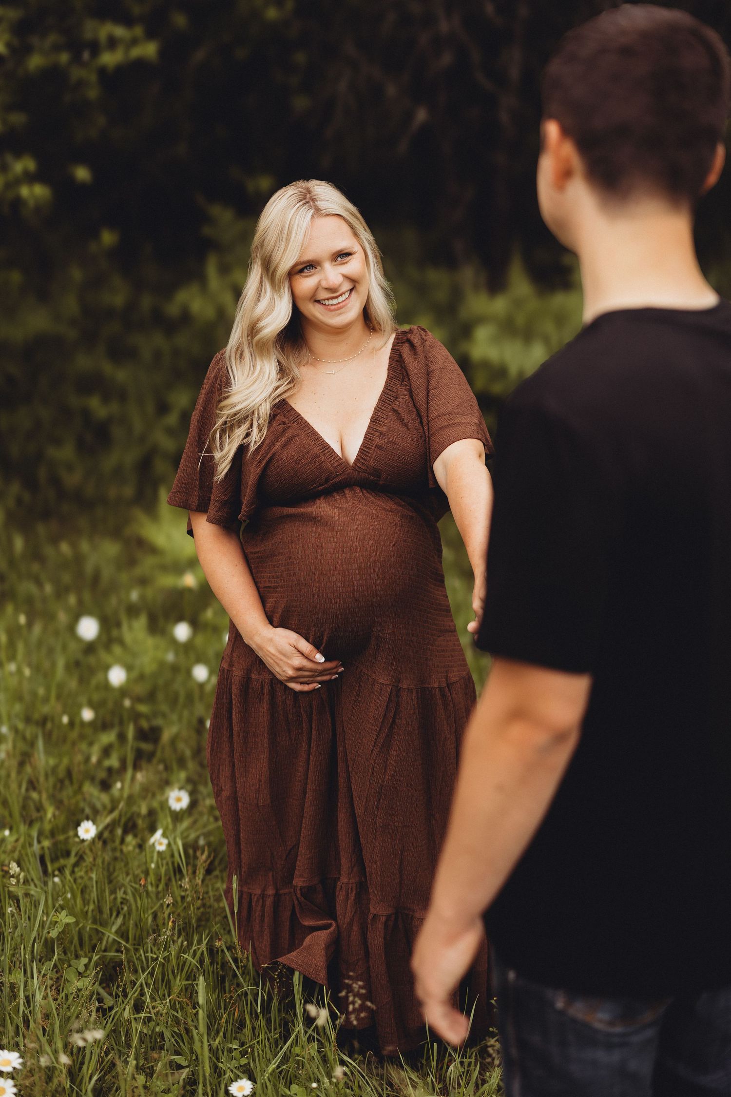 Smiling pregnant woman in brown dress holds baby bump in green field.