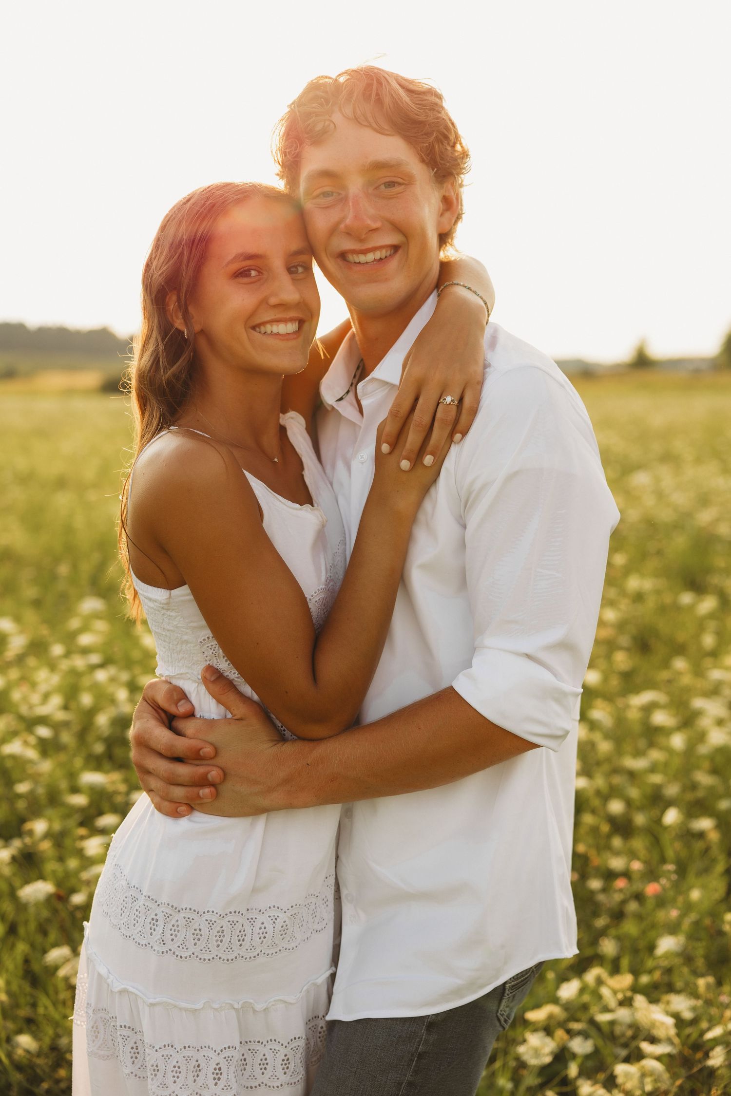 Happy young couple hugging and smiling in a sunlit flower field, wearing white outfits during golden hour.