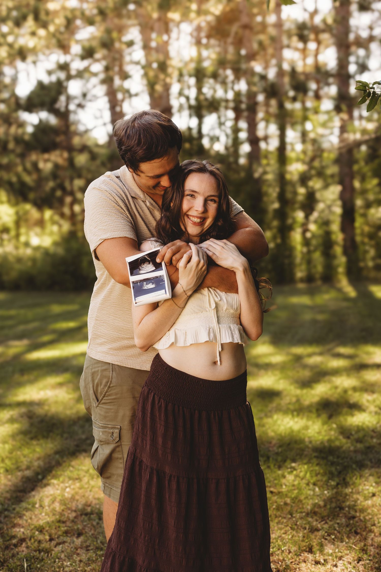 Happy pregnant couple embracing outdoors holding ultrasound photos in sunny forest setting.