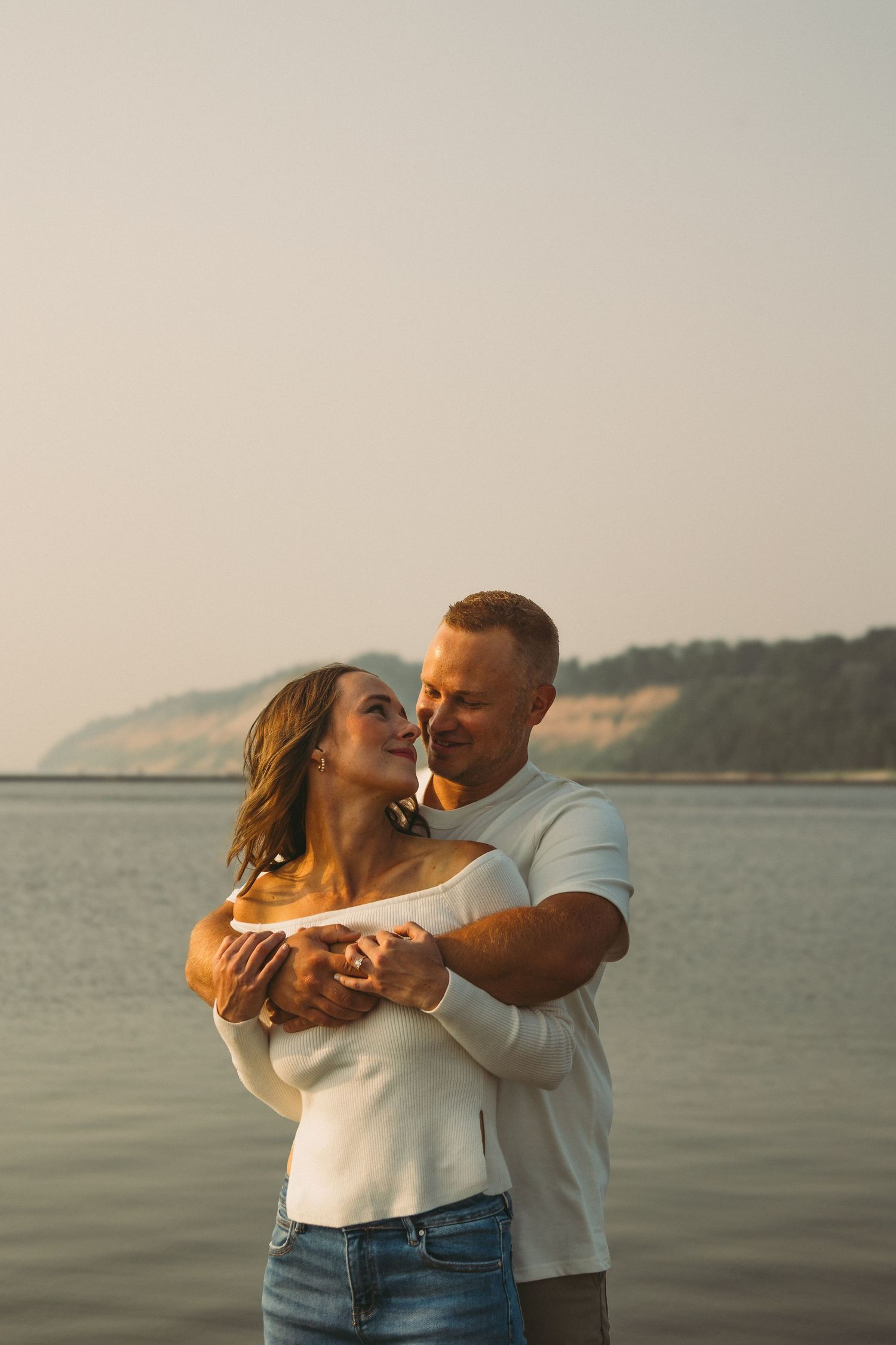 A couple embraces and smiles near a serene lake with scenic bluffs at golden hour.