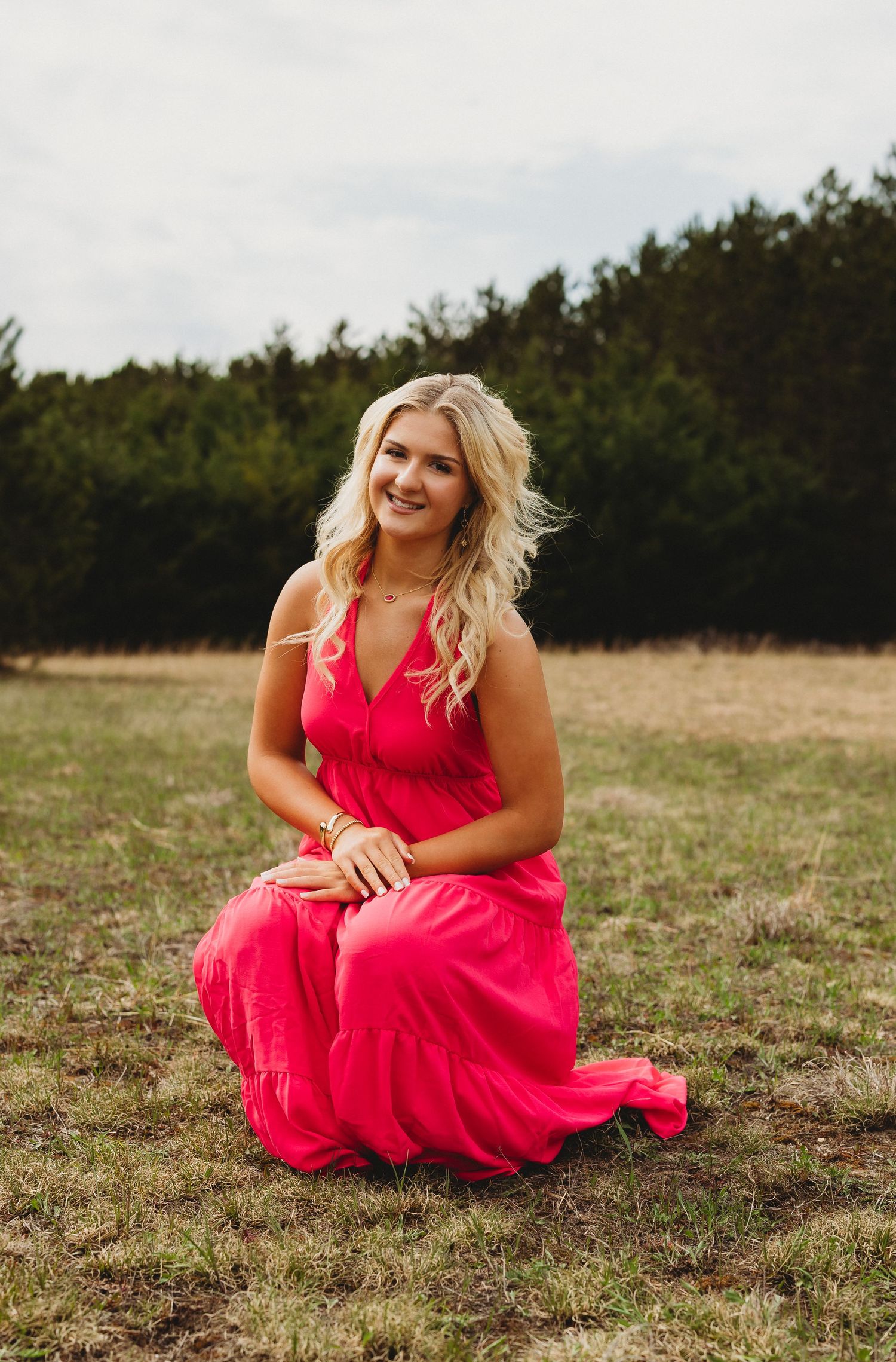 Blonde woman in a flowing pink maxi dress kneeling gracefully in a green field with trees in the background.