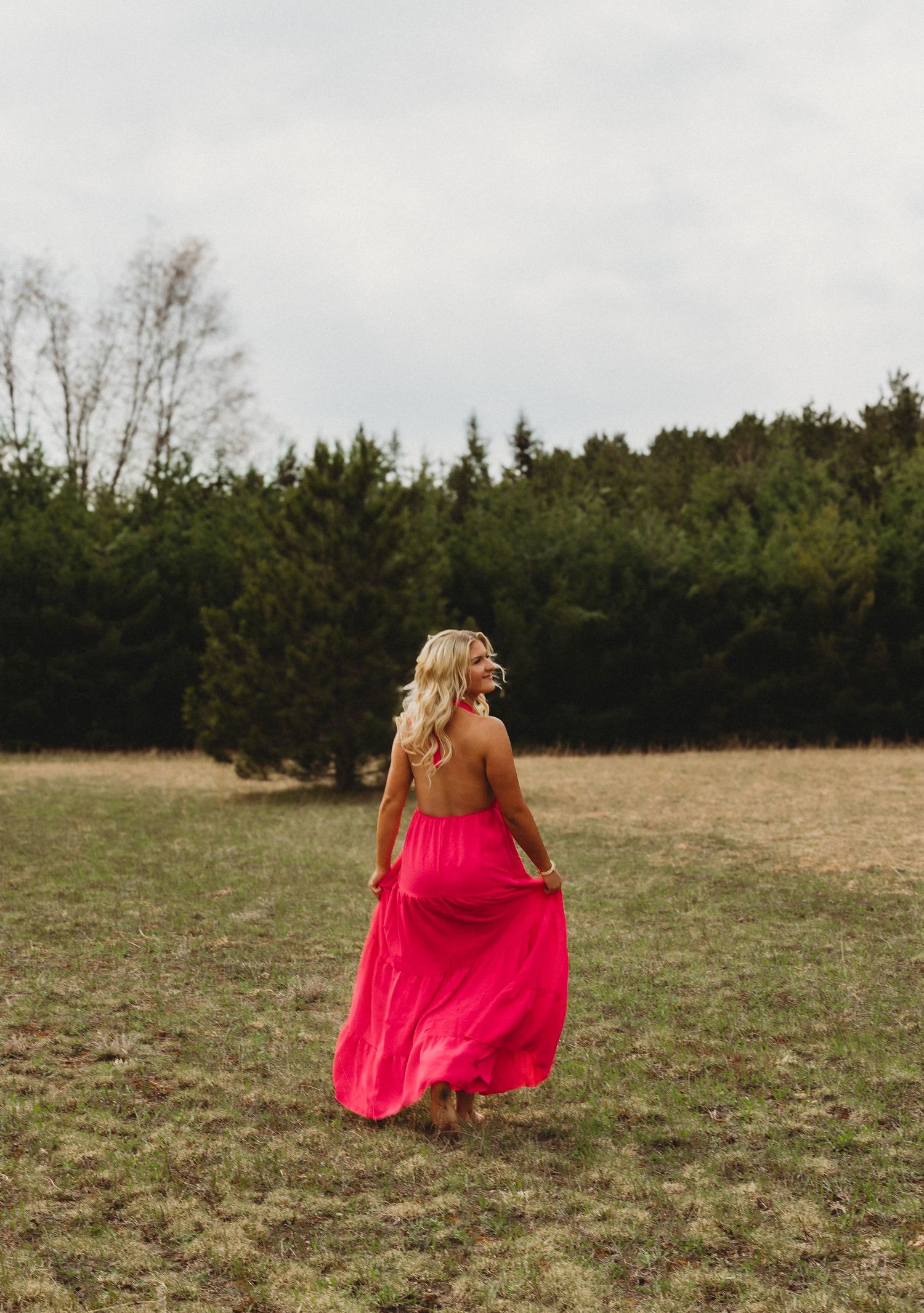 Blonde woman in a flowing pink halter maxi dress walking through a grassy field with evergreen trees behind her.