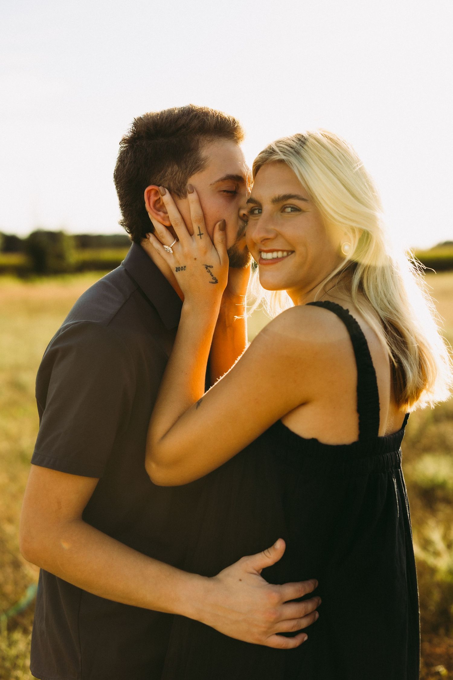 Smiling blonde woman in a black dress holds a man's face as he kisses her cheek in a golden sunlit field.