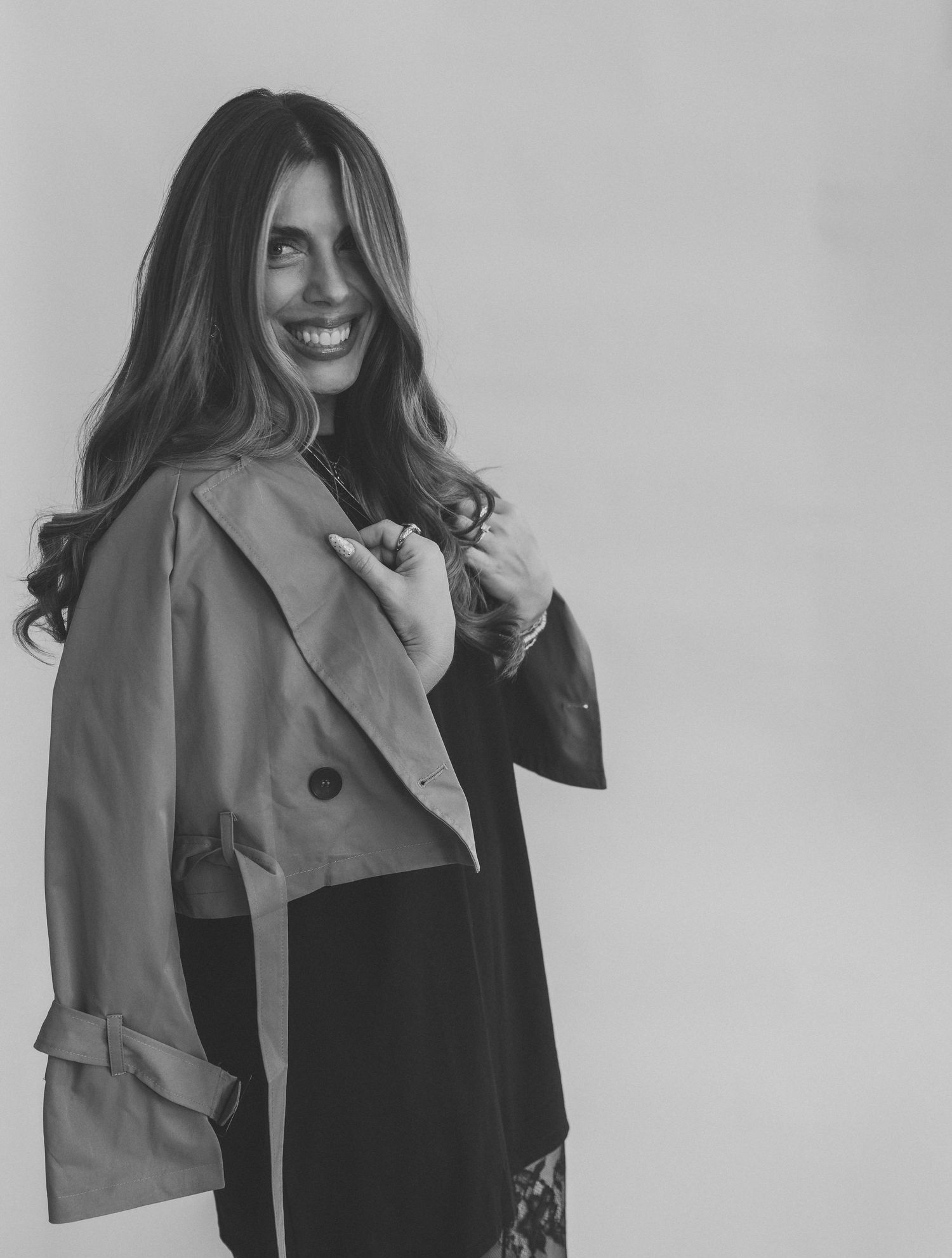 Black and white portrait of a smiling woman with long wavy hair wearing a cropped trench coat over a dark dress.