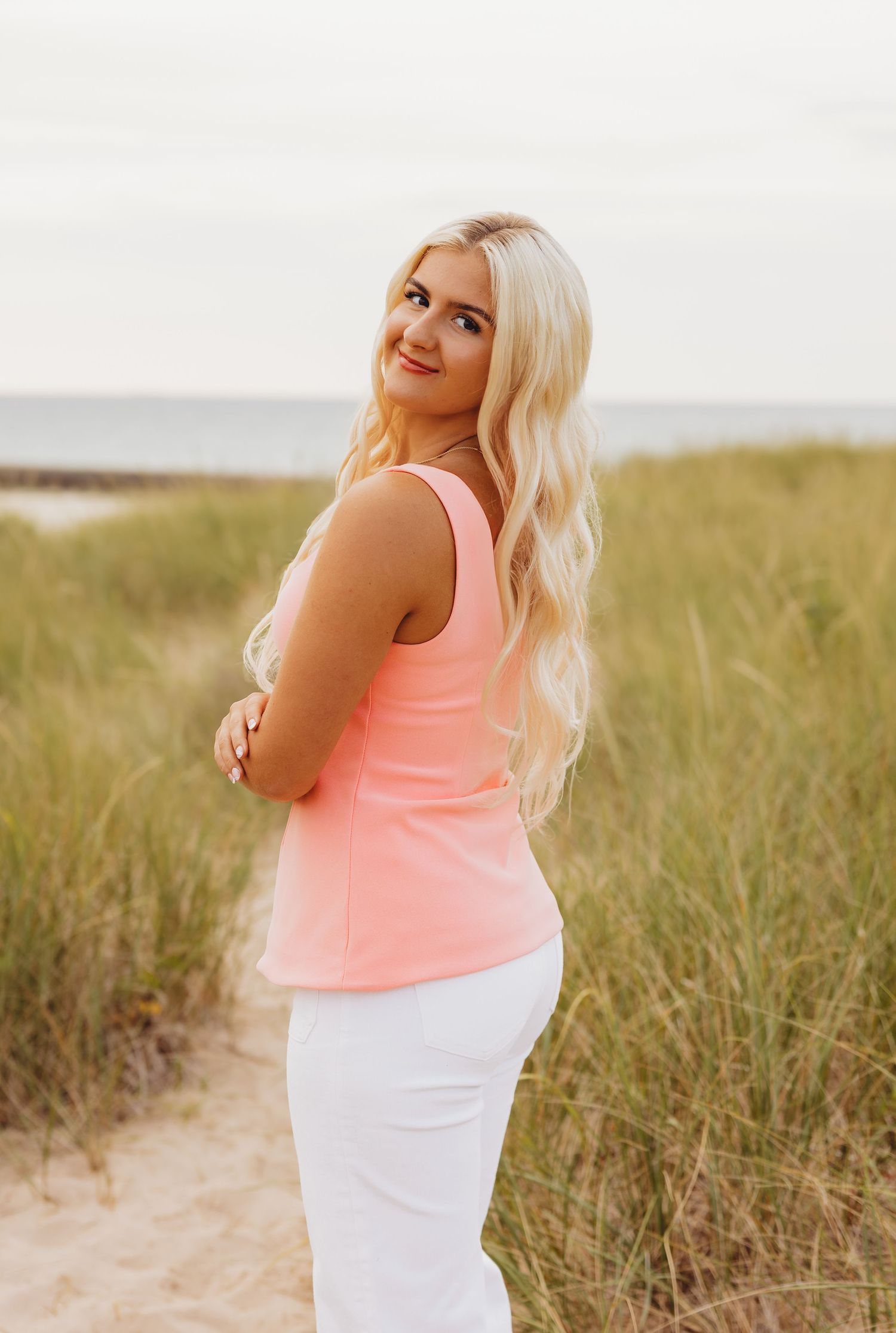 Smiling woman with long blonde hair in a coral tank top and white pants posing on a sandy path through beach grass near the water.
