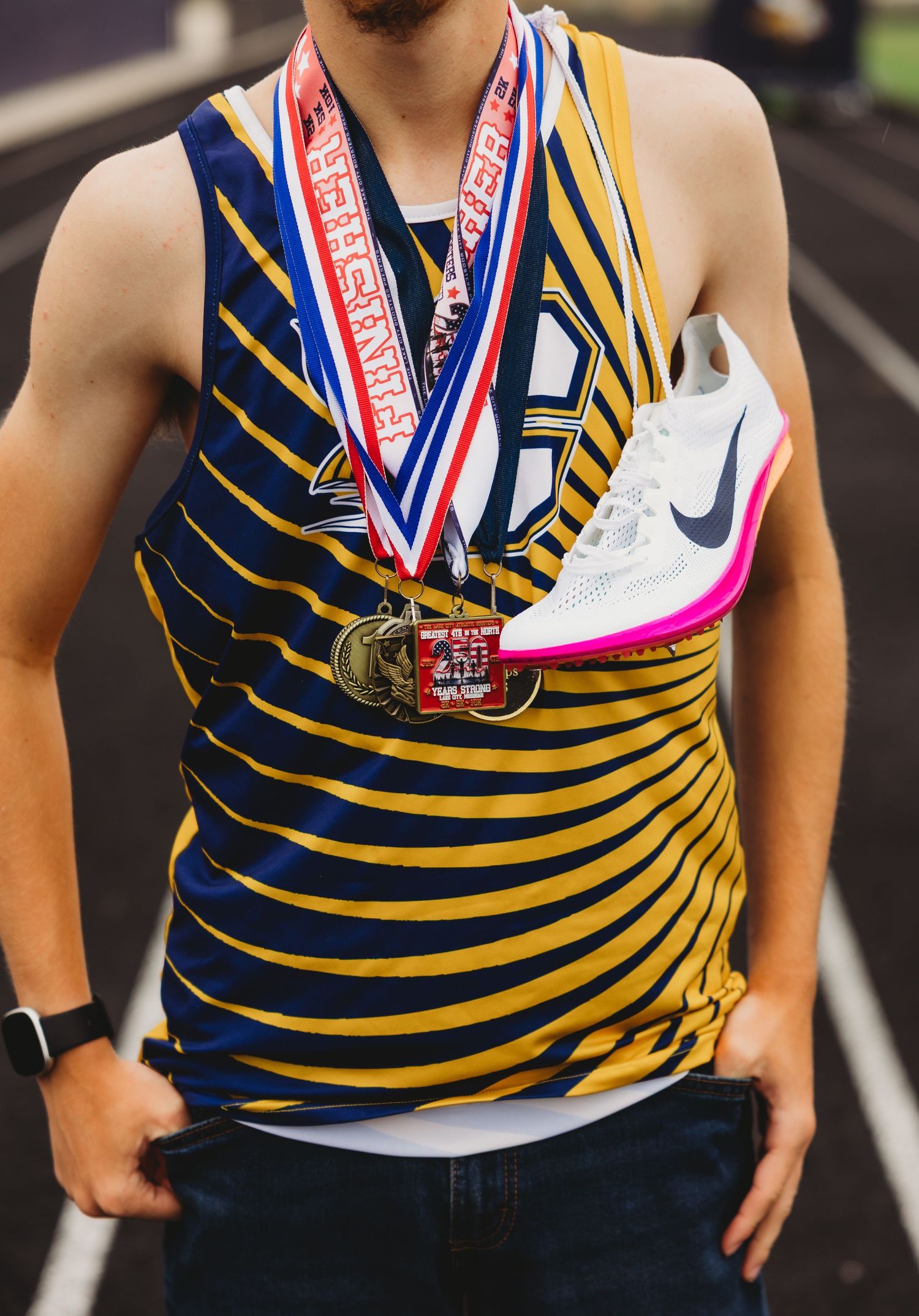 Track athlete in a blue and gold striped jersey wearing several medals and holding a white running spike with pink sole on the shoulder.