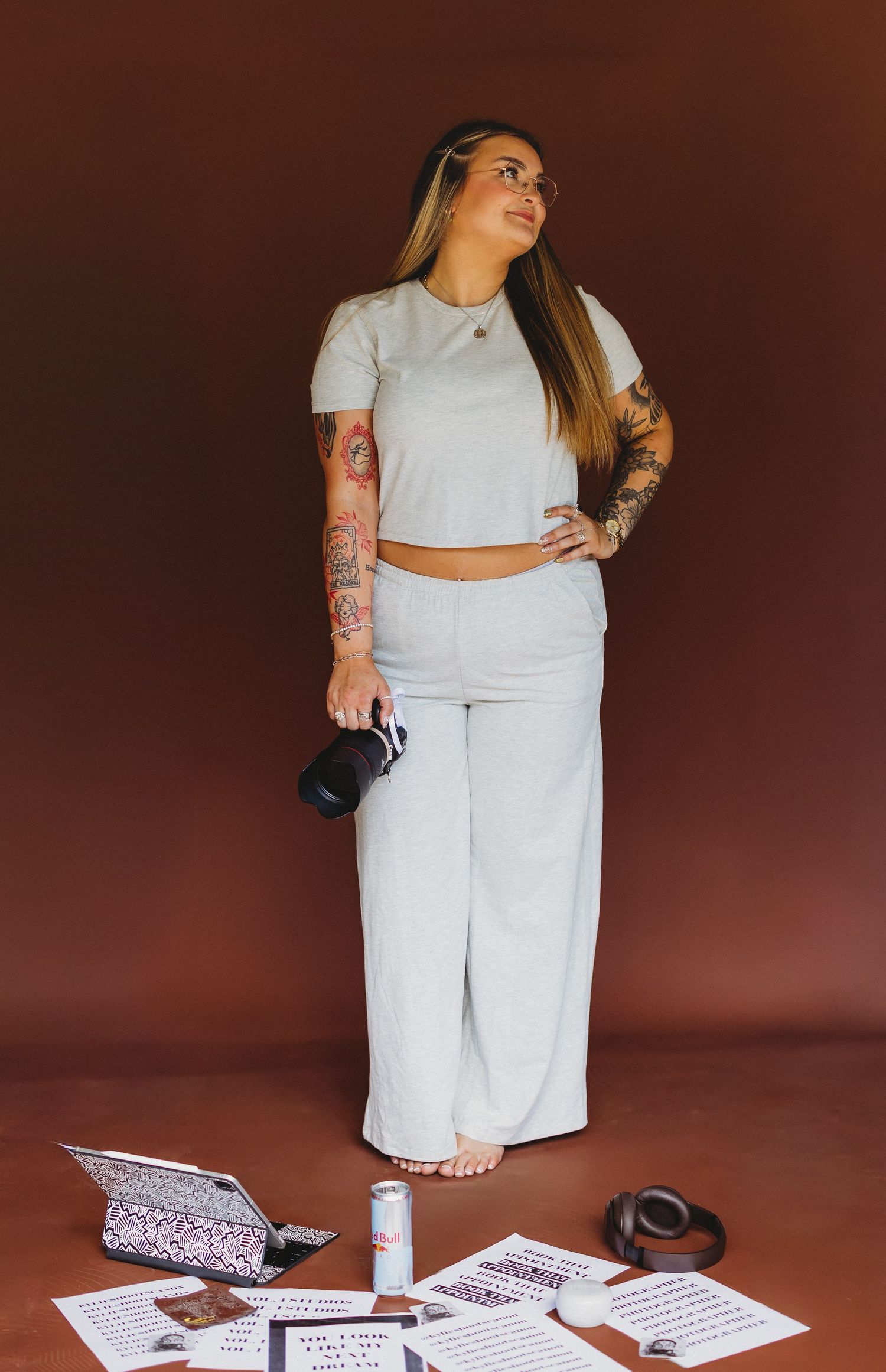 Tattooed woman in gray crop top and wide-leg pants holds camera, standing over papers and products on brown background.