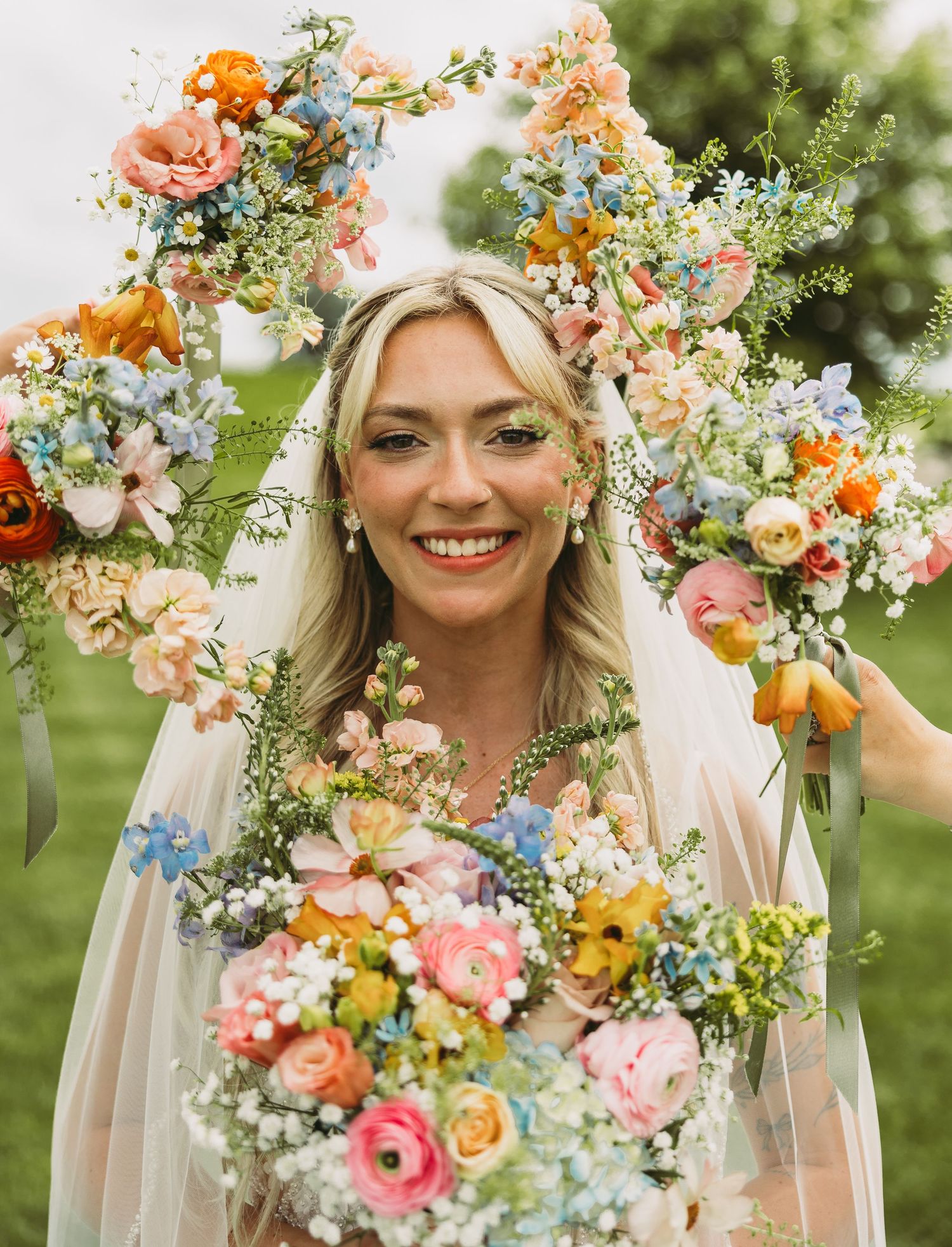 Joyful bride wearing a floral crown and veil surrounded by colorful spring flowers in an outdoor garden setting.