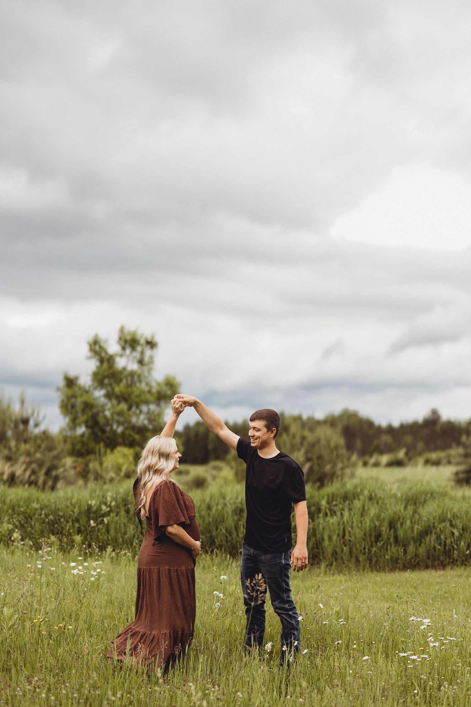 A couple dances together in a green meadow, the woman in a brown dress being twirled by a man in a black shirt.