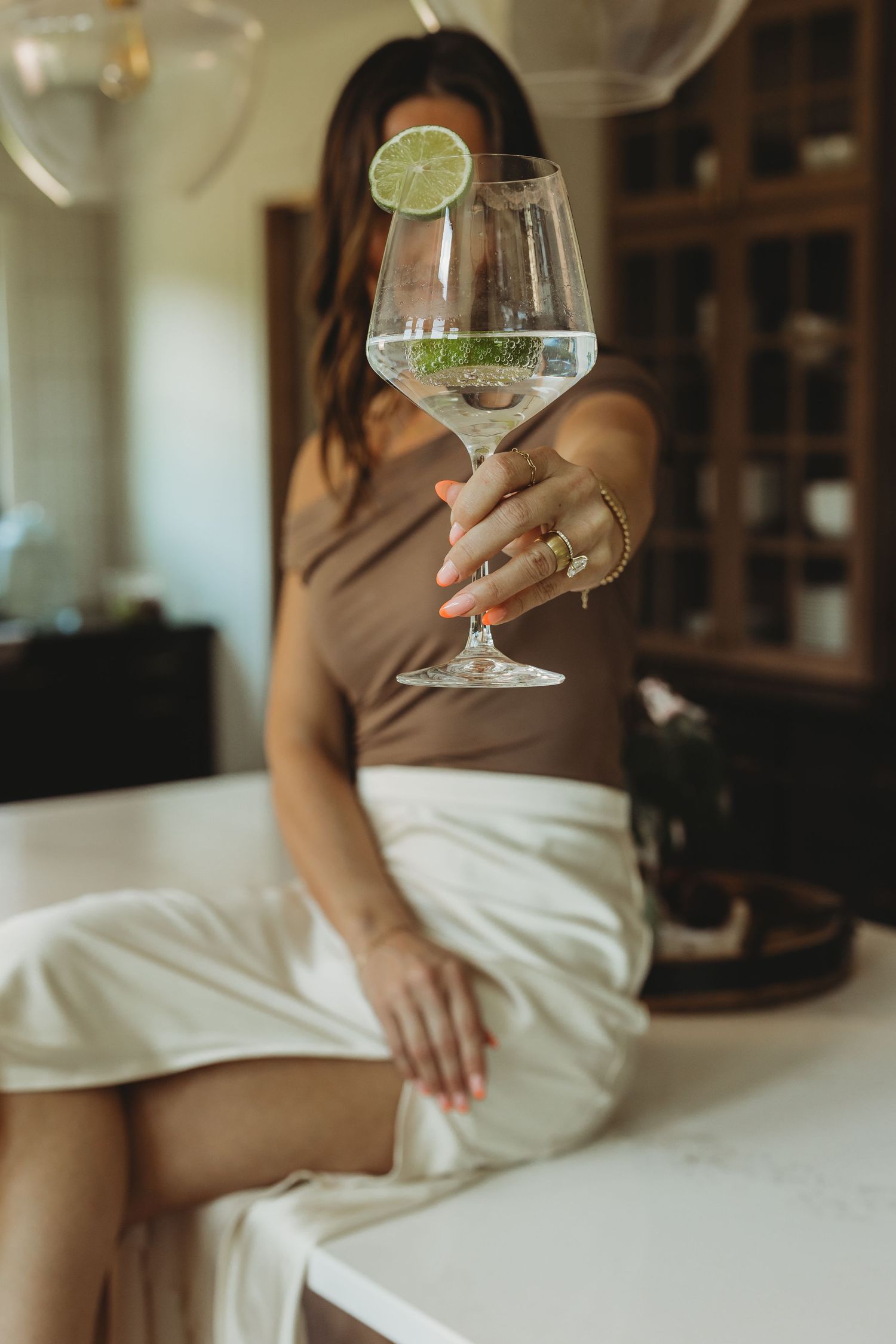 Woman in brown top holding a large wine glass with lime garnish while sitting on a white kitchen counter.