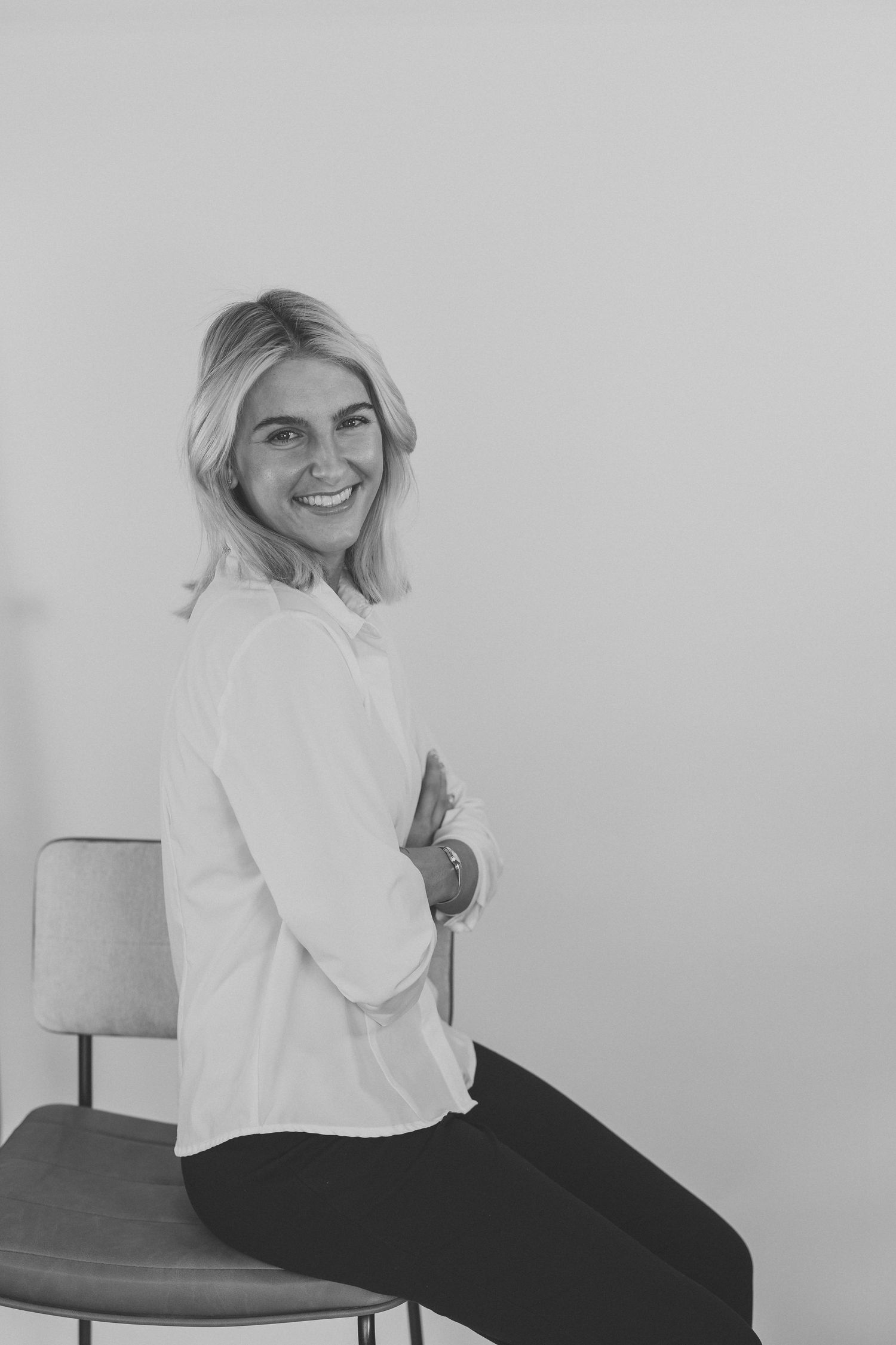 Smiling blonde woman in white shirt sitting on chair with arms crossed in black and white photo.