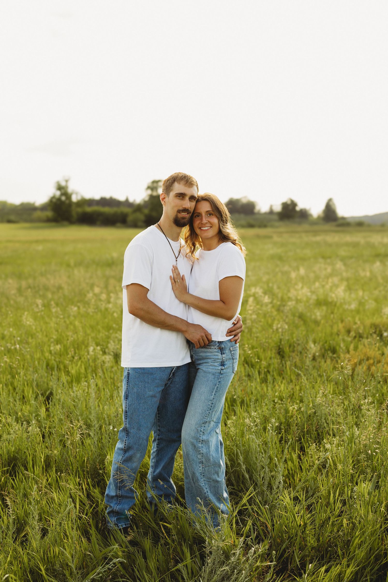 Smiling couple posing together in a lush green field during golden hour in matching white tees.
