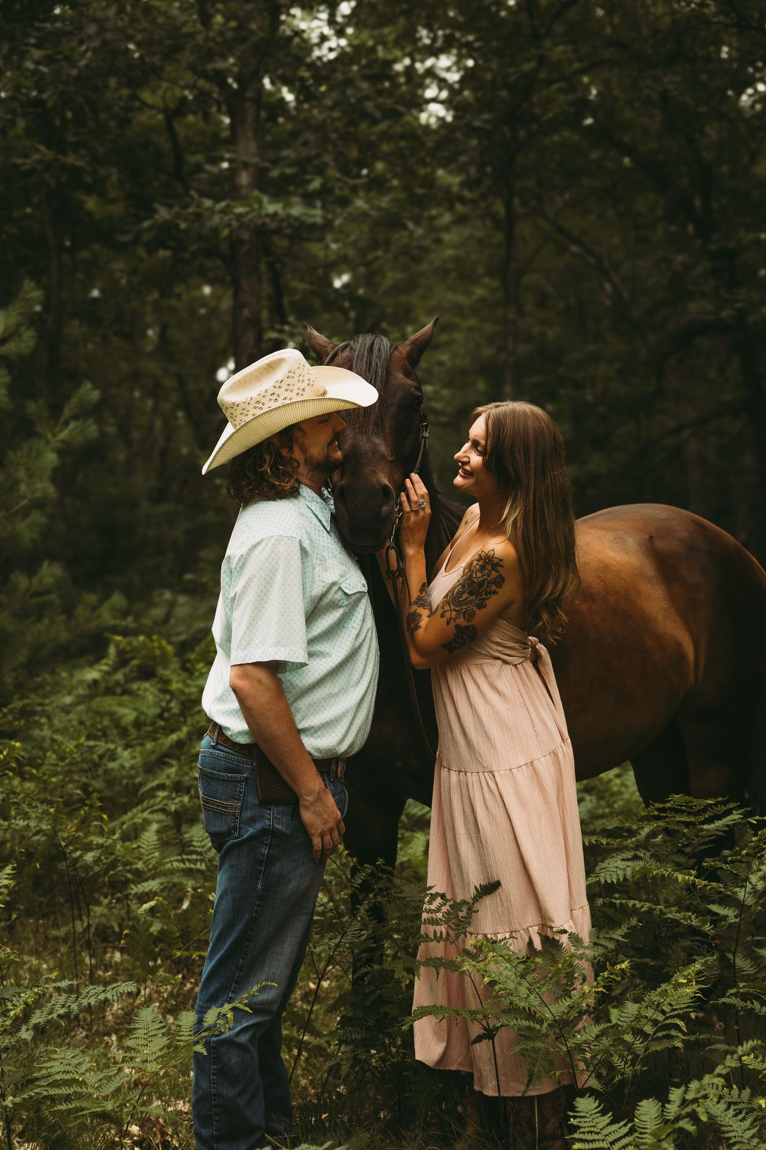 A couple shares a romantic moment with a brown horse in a lush forest, man in cowboy hat, woman in pink dress.