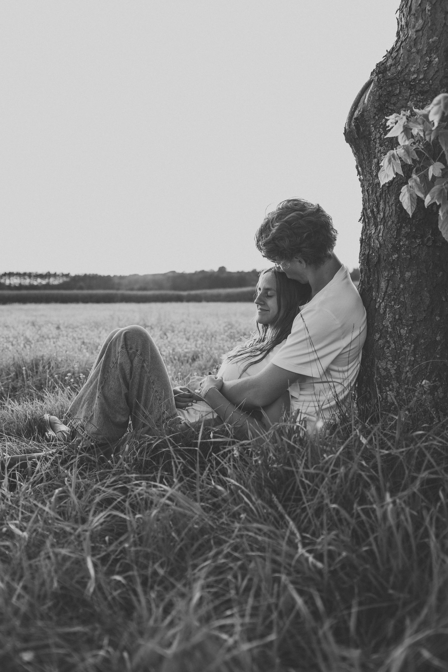 Black and white photo of a couple sitting together against a tree in a field, sharing a tender embrace.