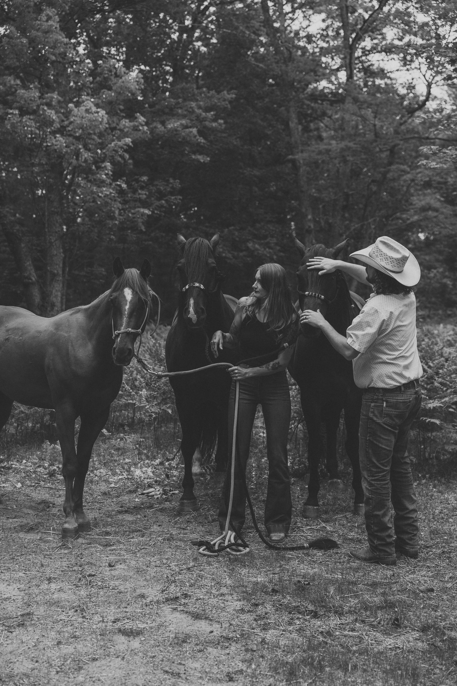 Black and white photo of two women with three horses in a wooded field, one wearing a cowboy hat.