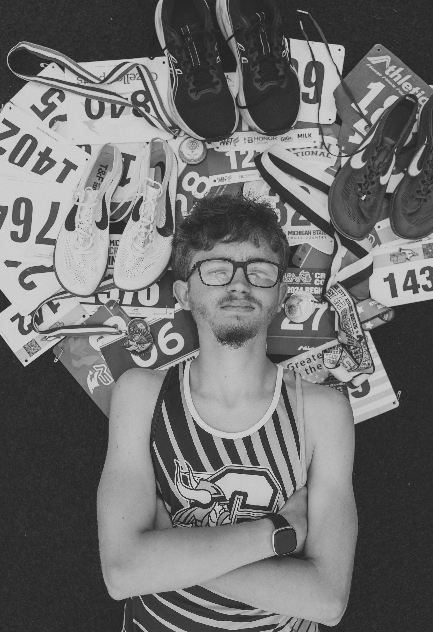 Black and white portrait of a young runner with glasses, arms crossed, lying among race bibs, running shoes, and medals.