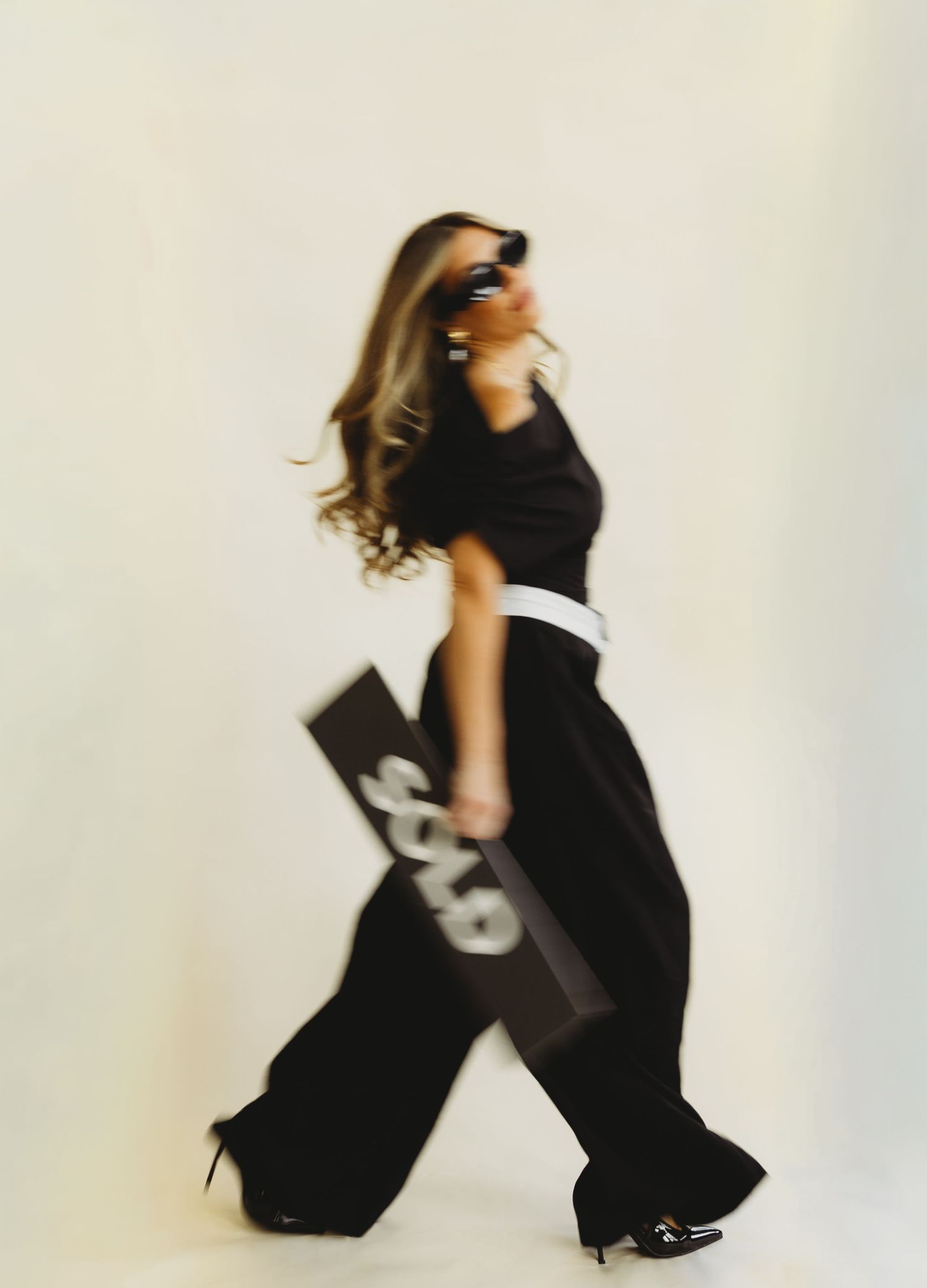 Woman in black wide-leg pants, white belt and sunglasses walks in motion blur while holding a black SOLD sign against a plain backdrop.