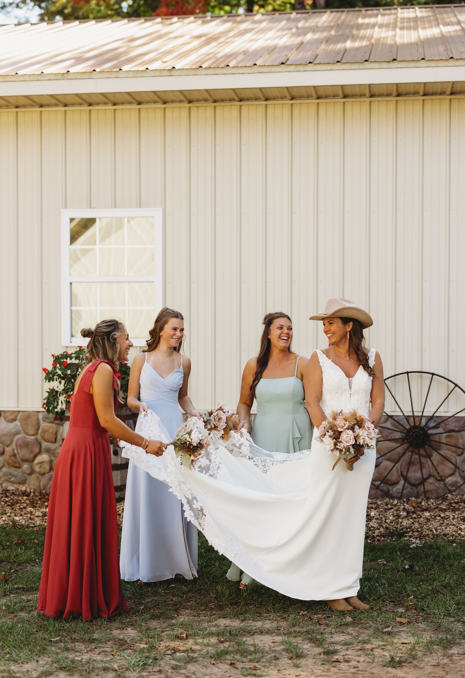 Bride in a white gown and cowboy hat holds her bouquet as three bridesmaids in red, blue and sage dresses laugh outside a rustic barn.