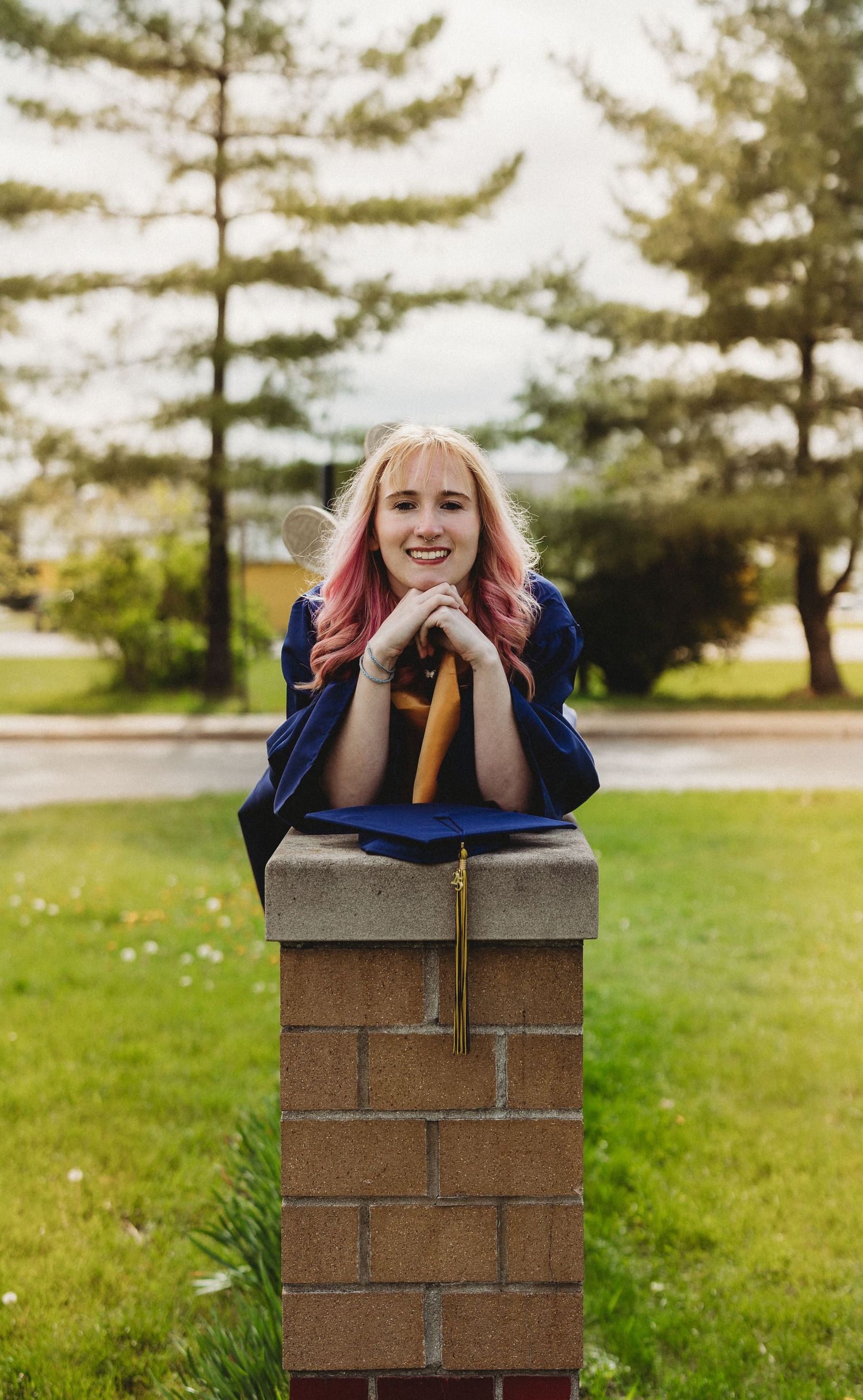 Graduate in blue cap and gown poses playfully at brick column in outdoor setting.