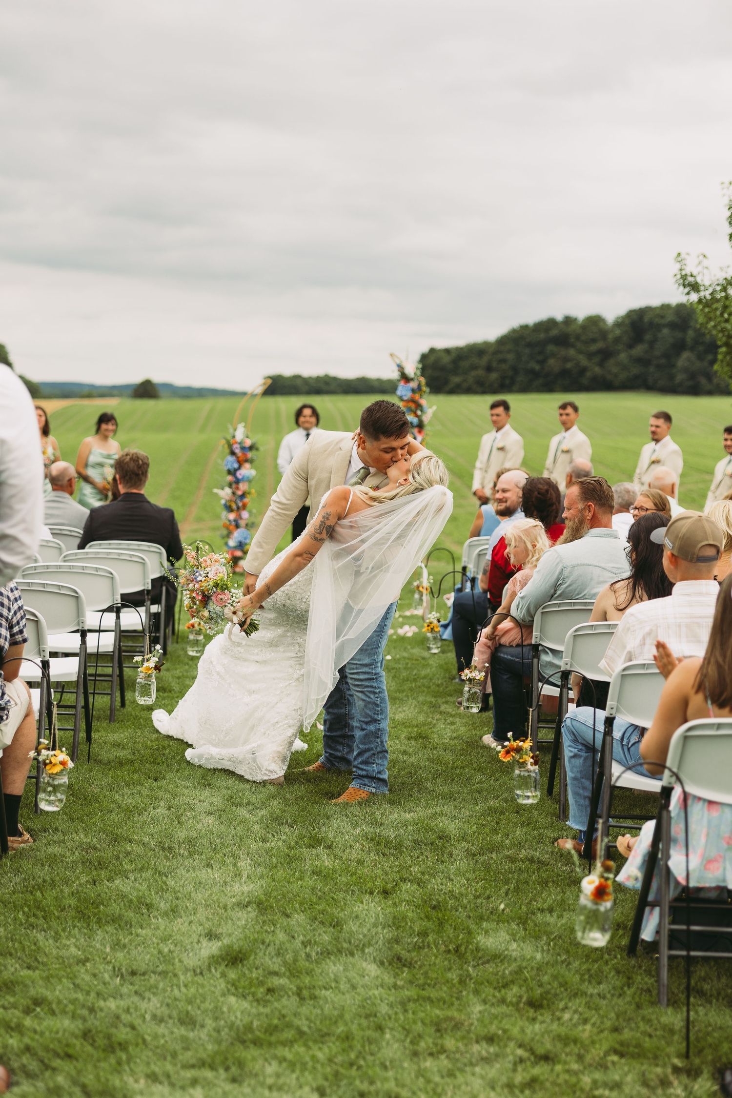 Wedding guests cheer as couple walks down outdoor aisle on grassy field.