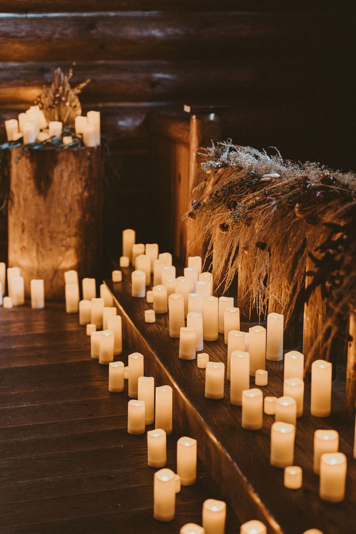 Glowing white candles line a dark wooden floor creating a romantic ambiance with dried pampas grass decorations.
