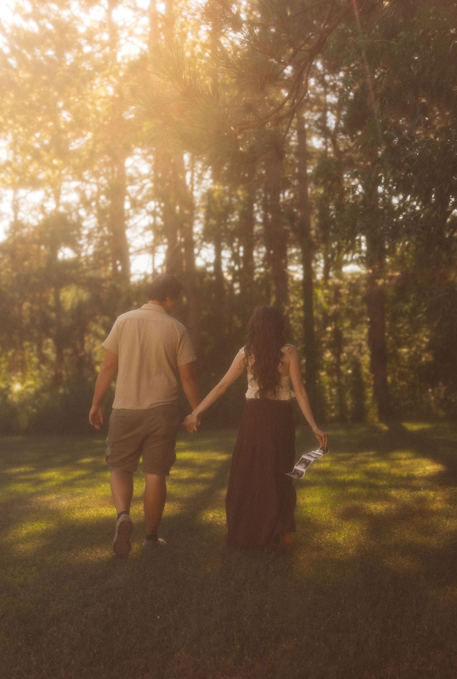 Couple holding hands walking through sunlit forest park on grass at golden hour sunset.