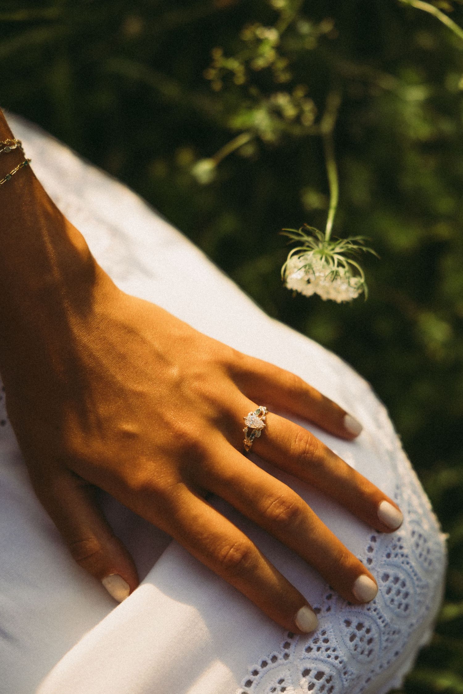 Close-up of a woman's hand wearing a diamond engagement ring, resting on a white lace dress outdoors.