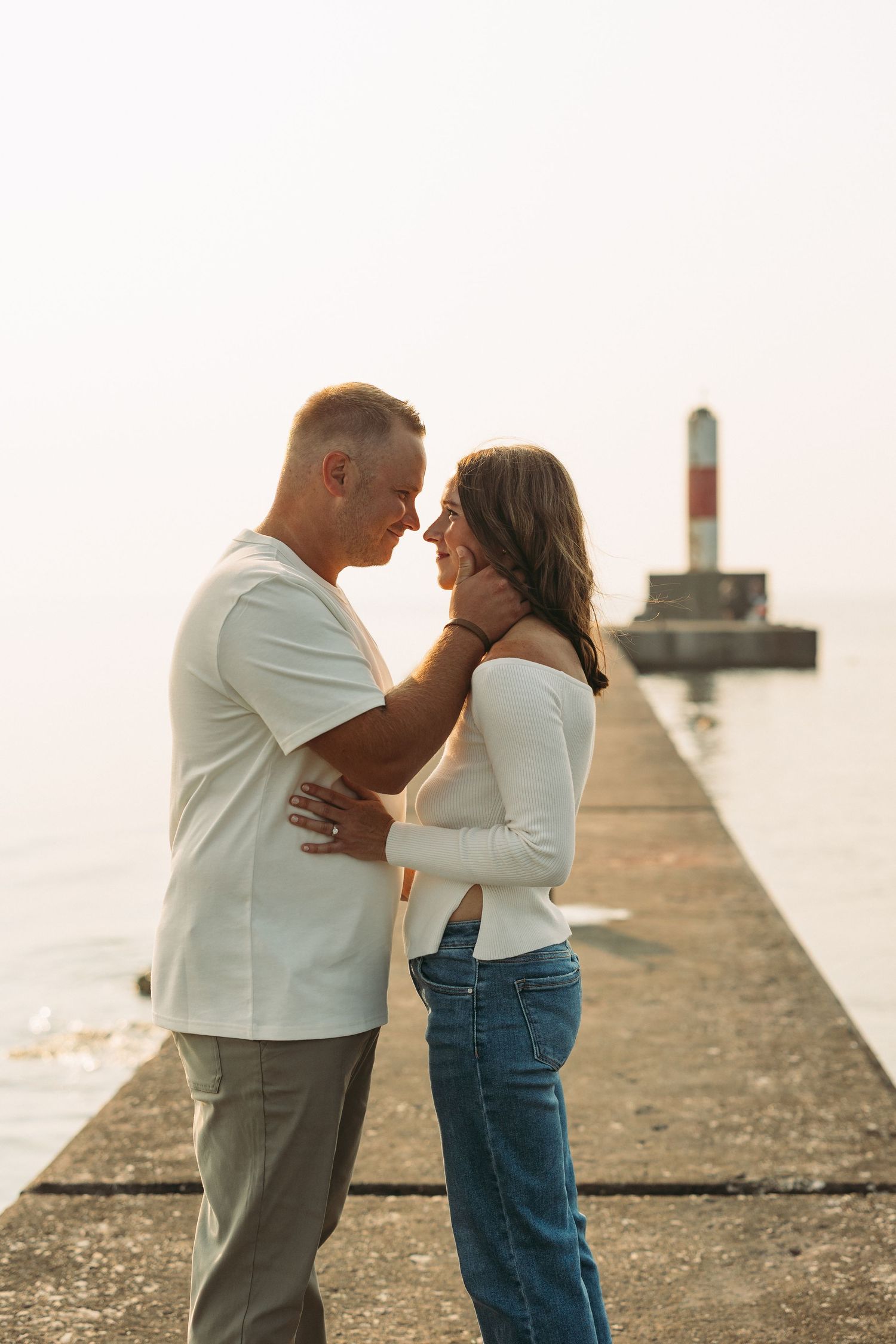 Romantic couple embracing on a waterfront pier at sunset, with a lighthouse in the background.