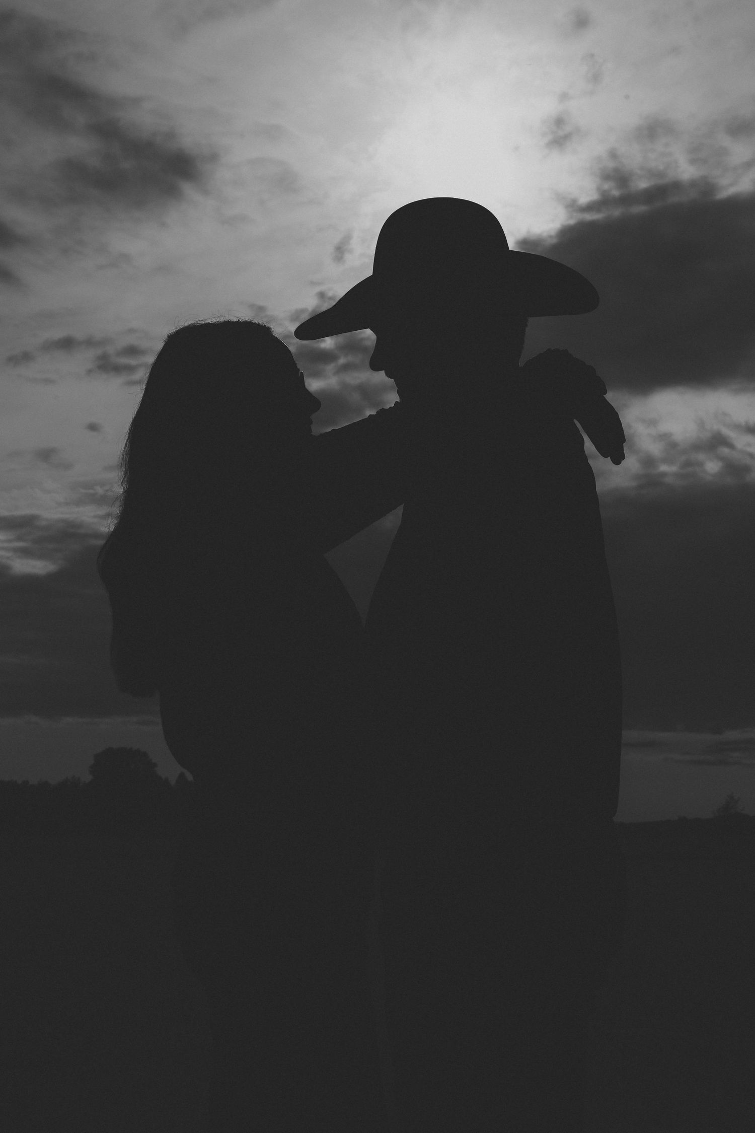 Black and white silhouette of a couple sharing a romantic moment under a dramatic cloudy sky at sunset.