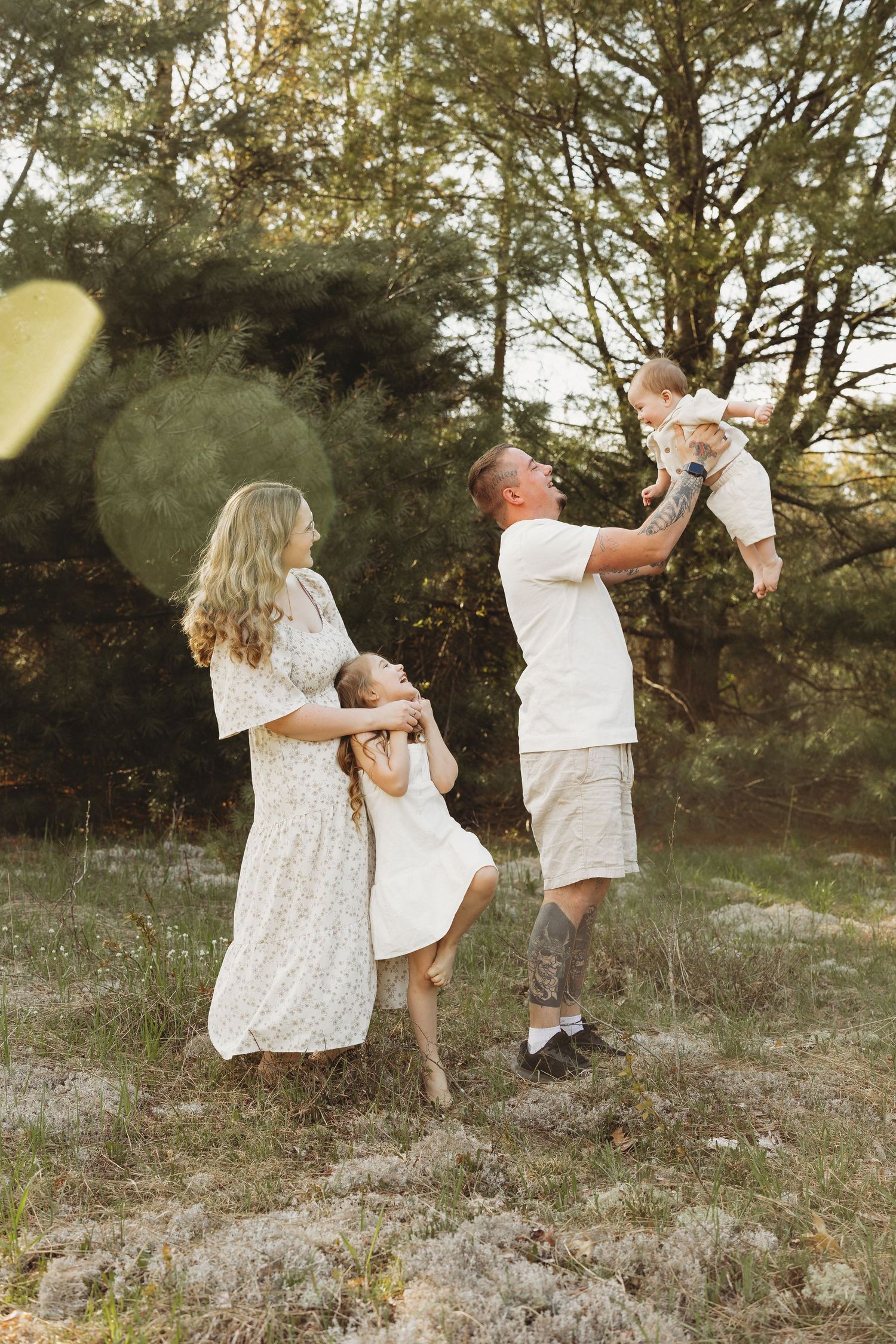 A joyful family of four plays outdoors in a sunlit forest, with parents in white outfits lifting and hugging their young children.