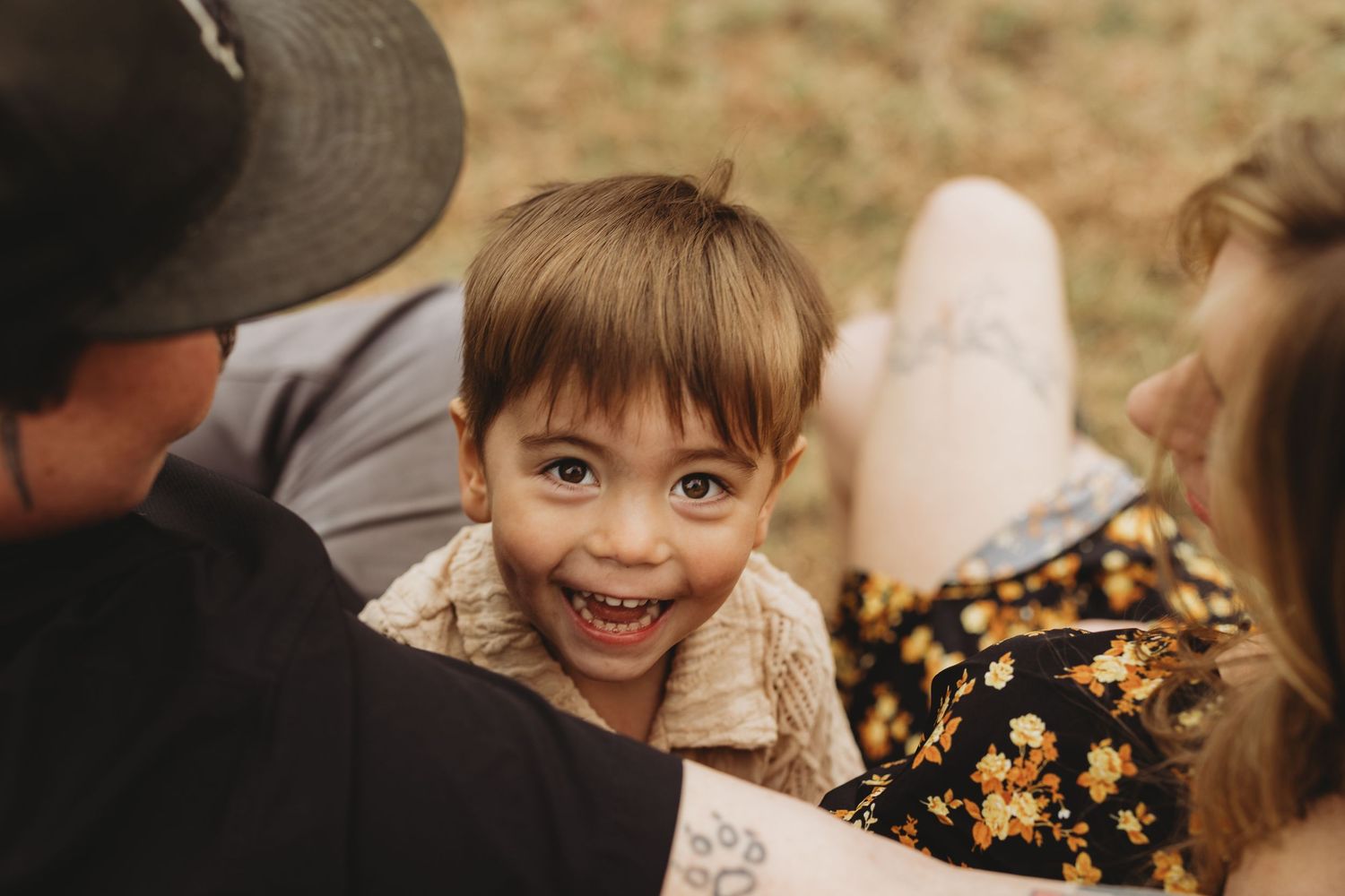 A joyful toddler smiles brightly while sitting between two adults outdoors in a warm, natural setting.