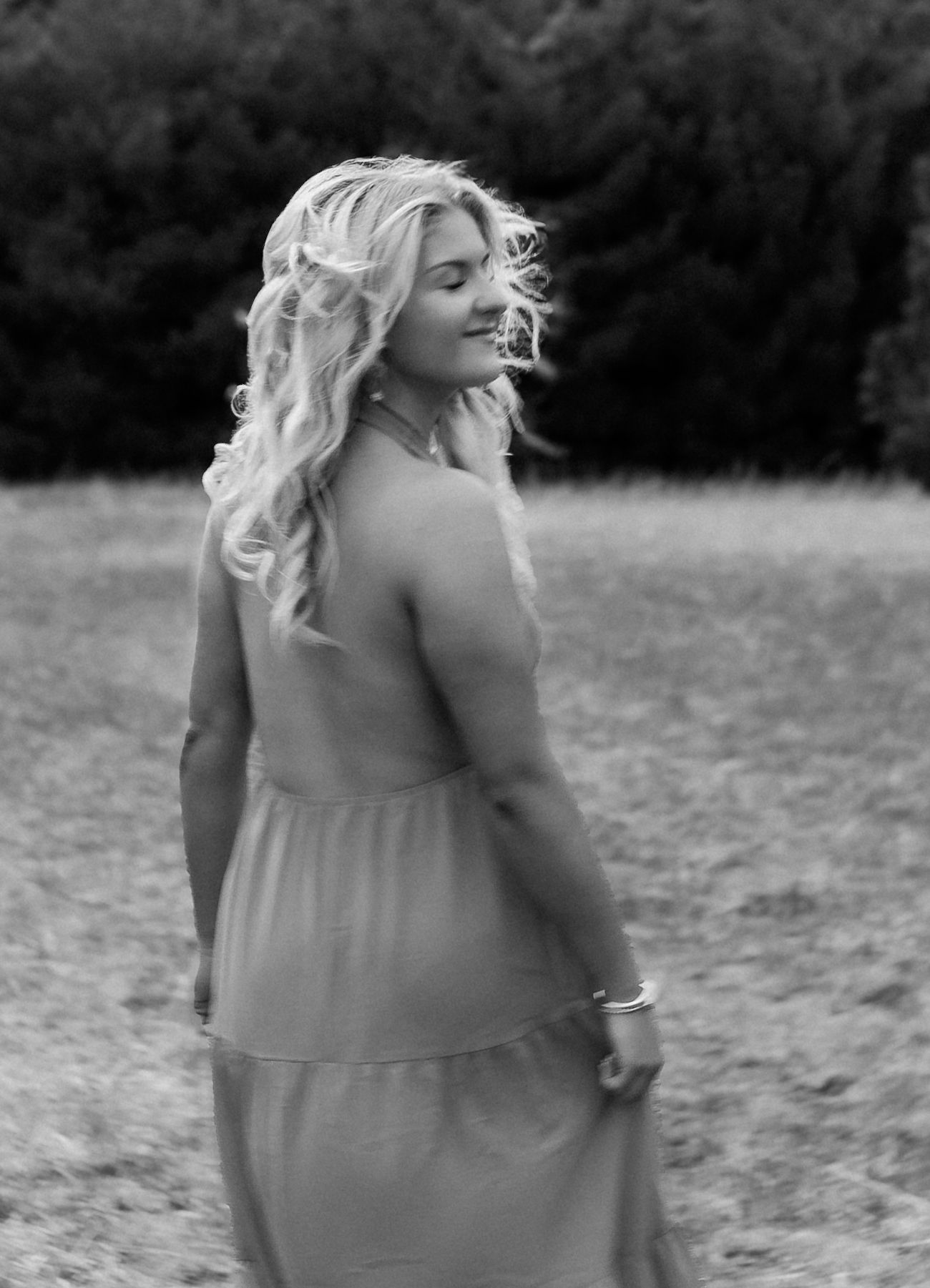 Black and white photo of a young blonde woman in a flowing dress, smiling over her shoulder in an open field.