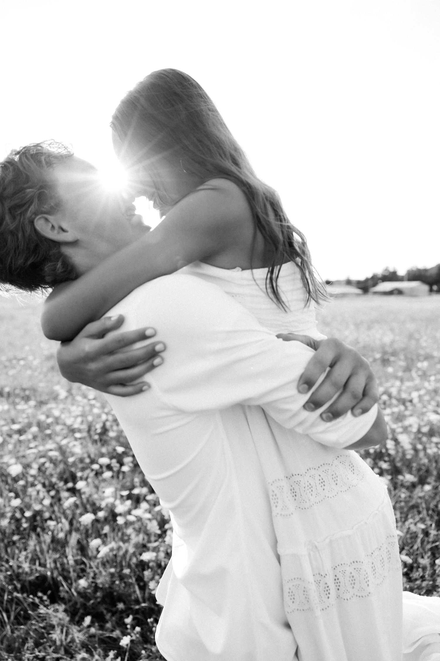Black and white photo of a couple embracing in a sunlit flower field, woman in white dress lifted by her partner.