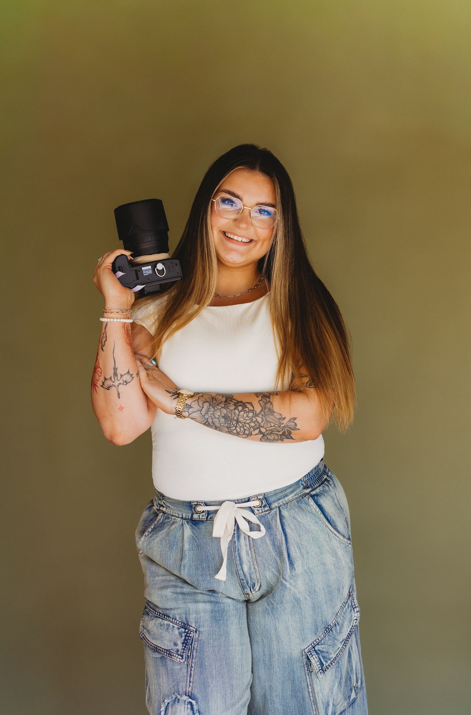 Smiling woman with tattoos holding a camera, wearing glasses, white top, and wide-leg jeans against a neutral background.