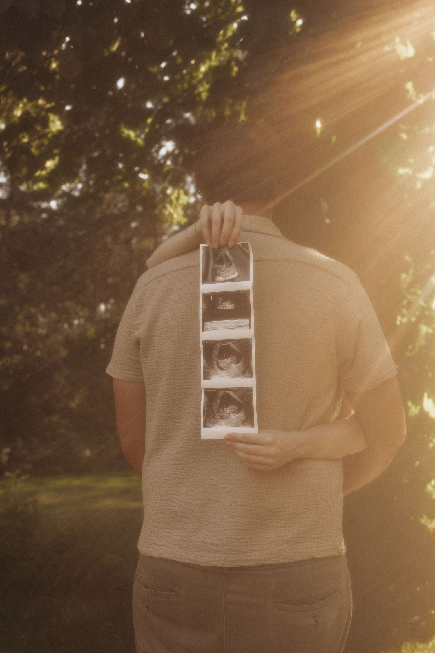 Couple embracing outdoors holding ultrasound photos in golden sunlight, pregnancy announcement in a lush green garden.