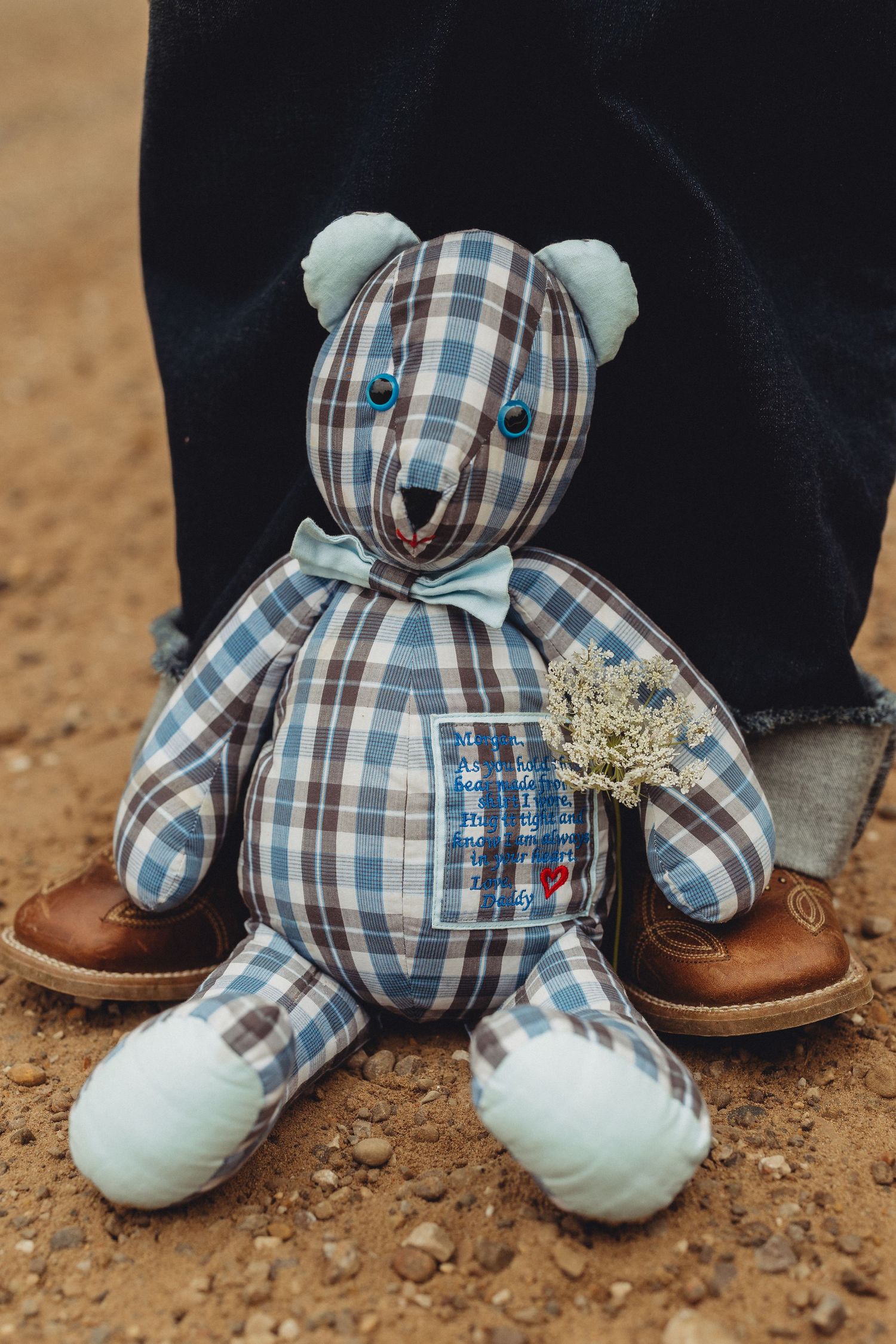 Handmade plaid teddy bear with bow tie sitting on gravel path near cowboy boots.