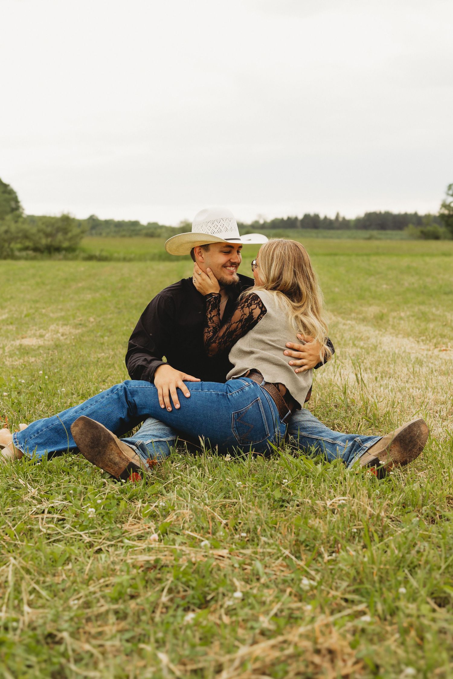 A couple sits romantically in a green field, the man wearing a cowboy hat and jeans, as the woman leans in for a kiss.