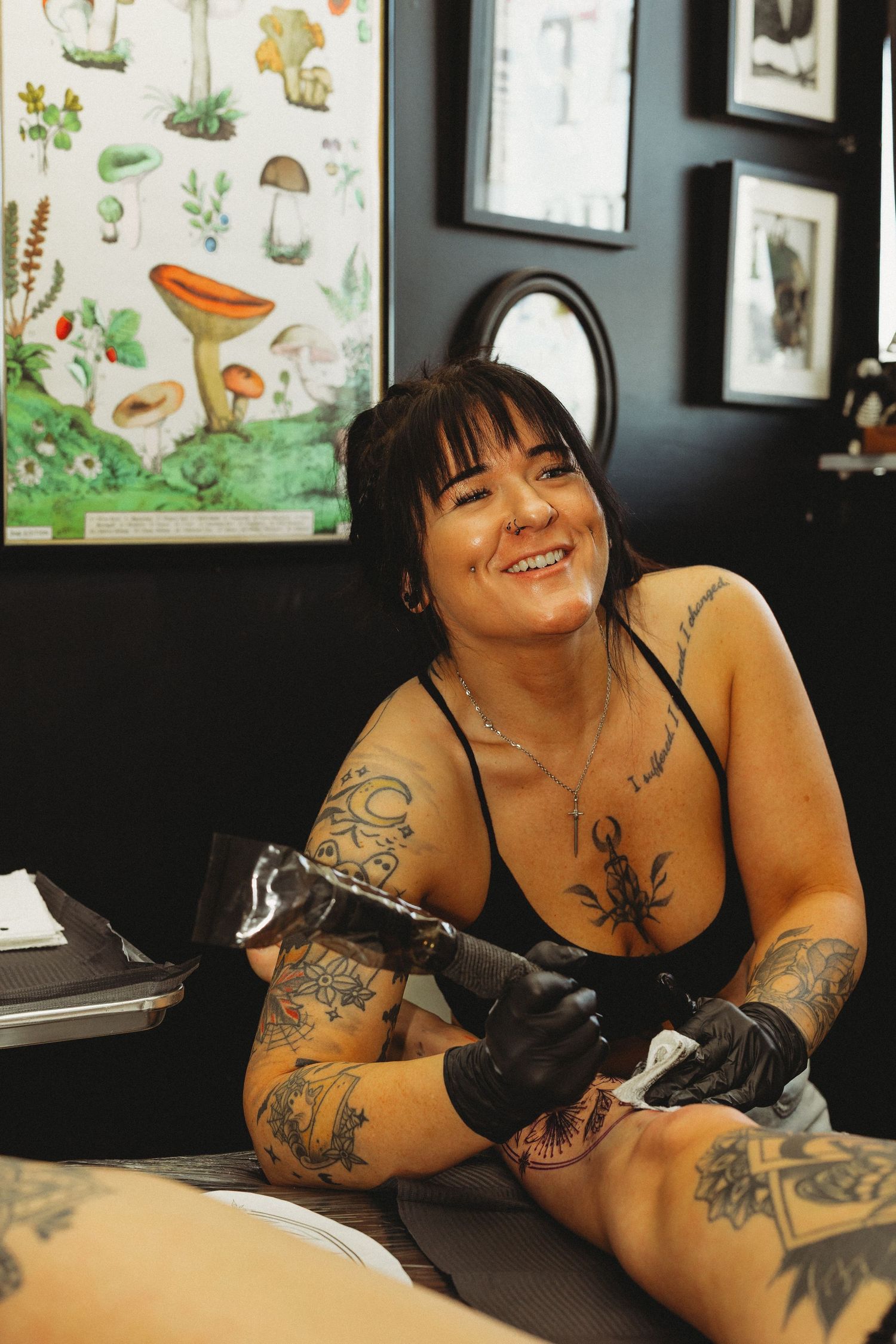 Smiling female tattoo artist with sleeve tattoos works on a client in a decorated studio with mushroom poster.