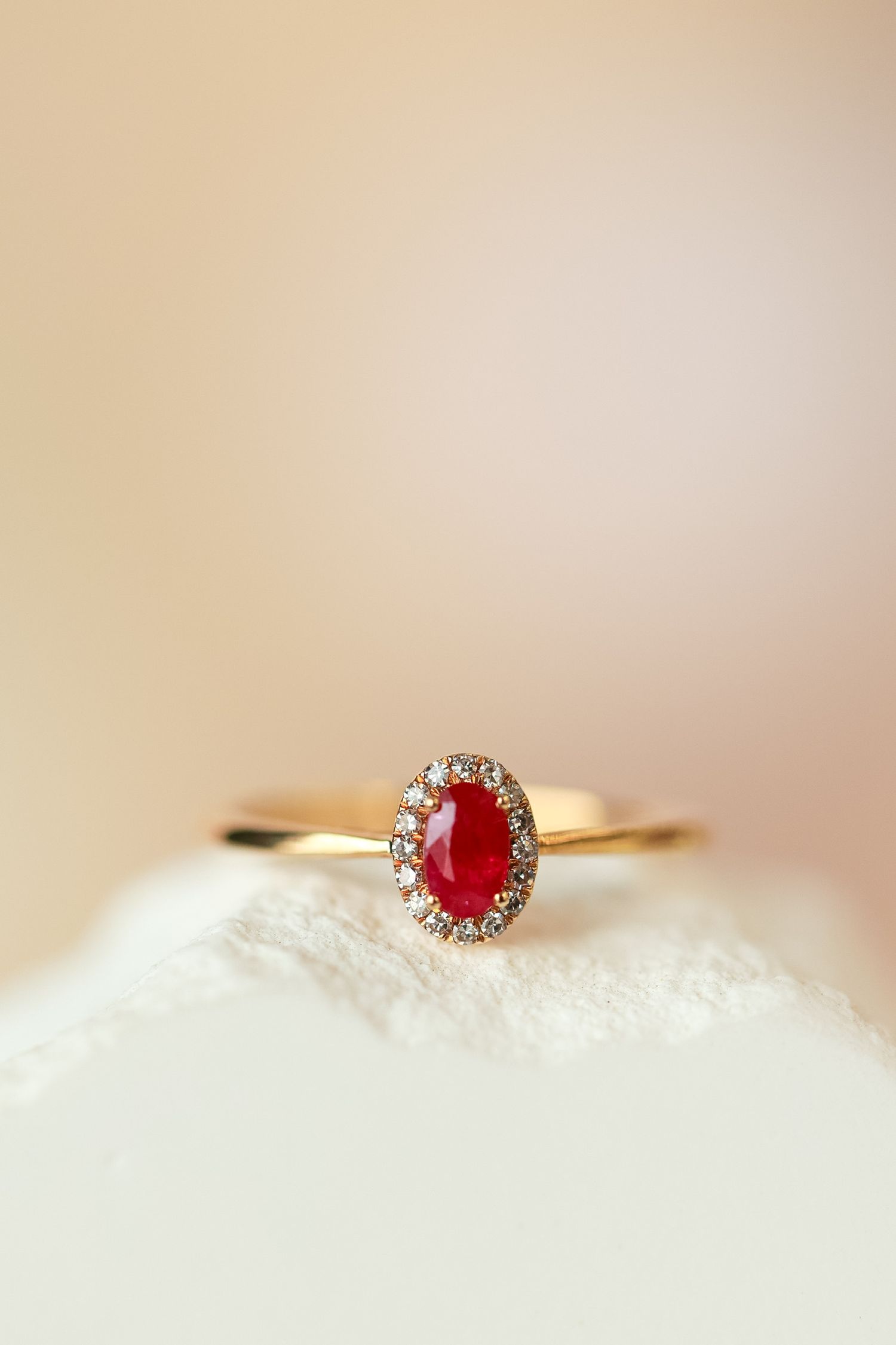 Delicate gold ring with small ruby and diamond halo on white textured surface.