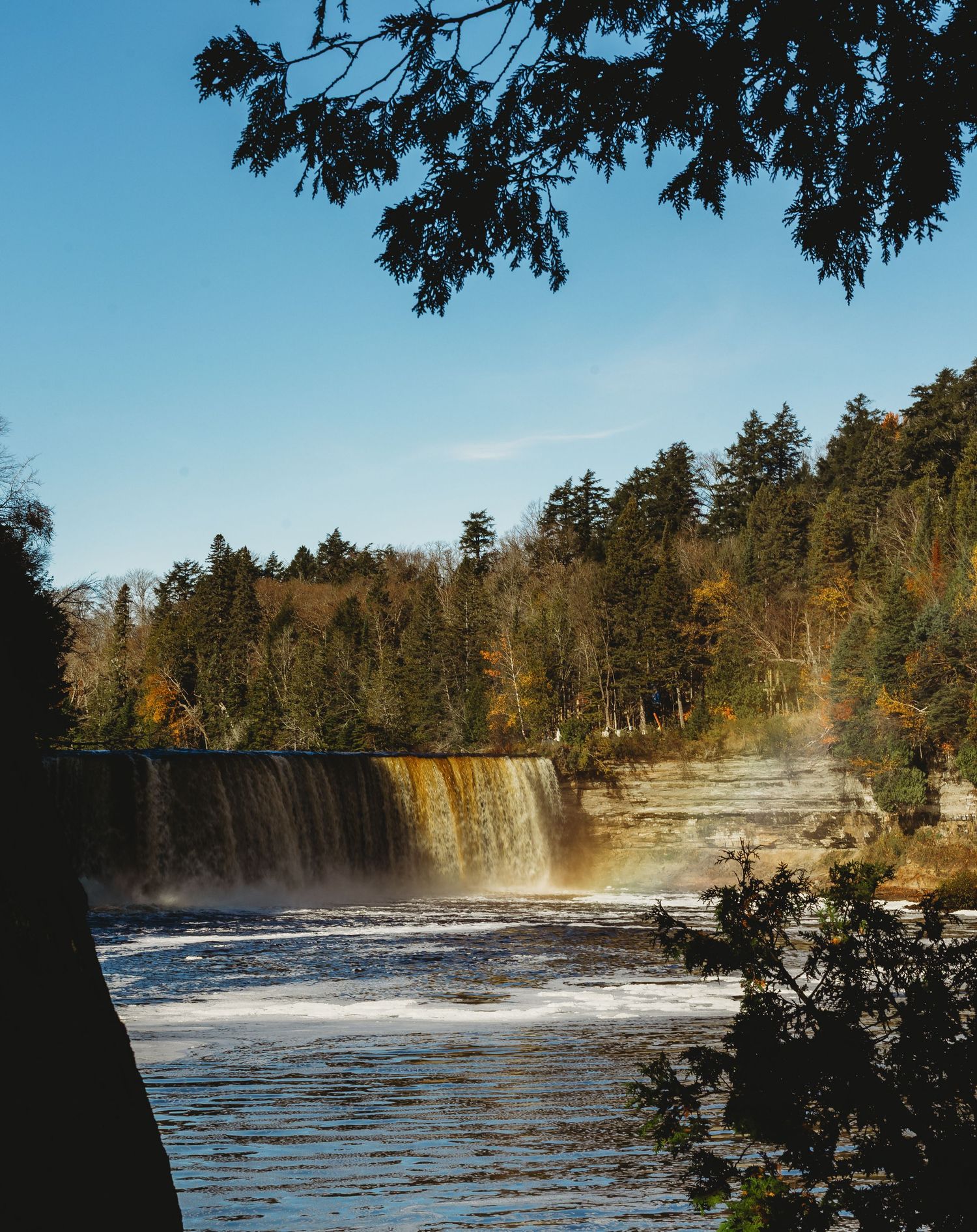 Scenic waterfall with rainbow mist and golden autumn trees under bright blue skies.