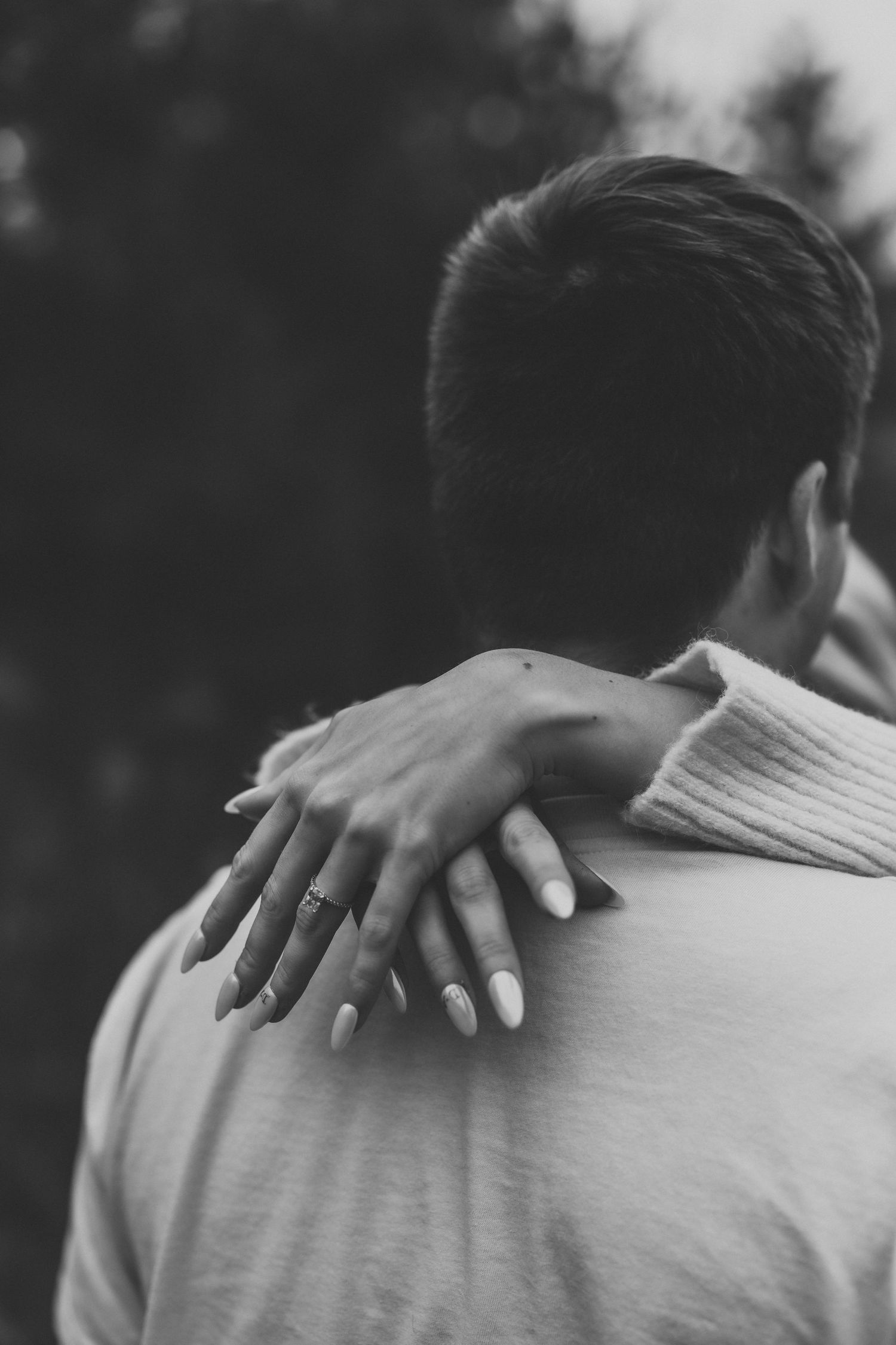 A close-up black and white photo showing hands with an engagement ring embracing a shoulder in an intimate moment.