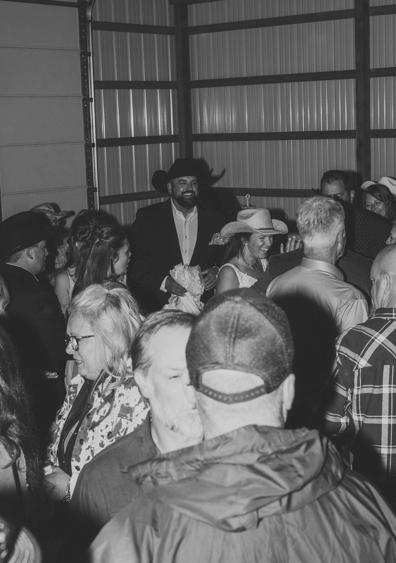Black and white photo of wedding guests in cowboy hats and casual attire mingling inside a rustic metal barn venue.