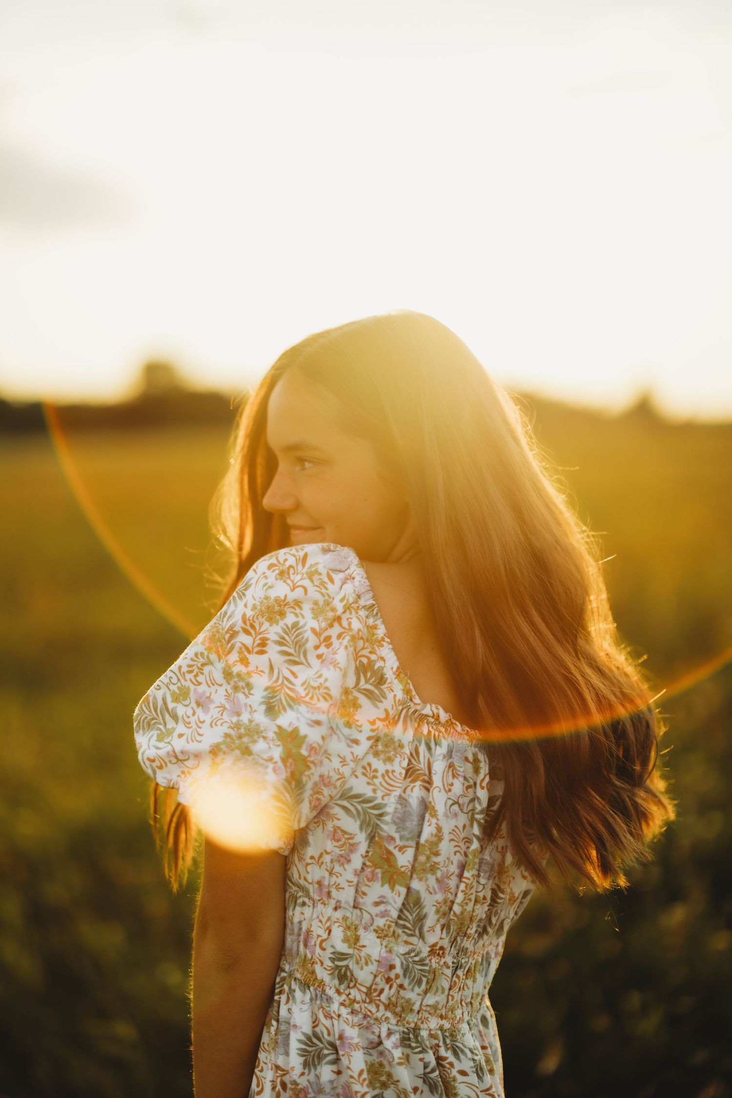 Young woman with long auburn hair in a floral dress glances over her shoulder in a golden field at sunset with warm lens flare.